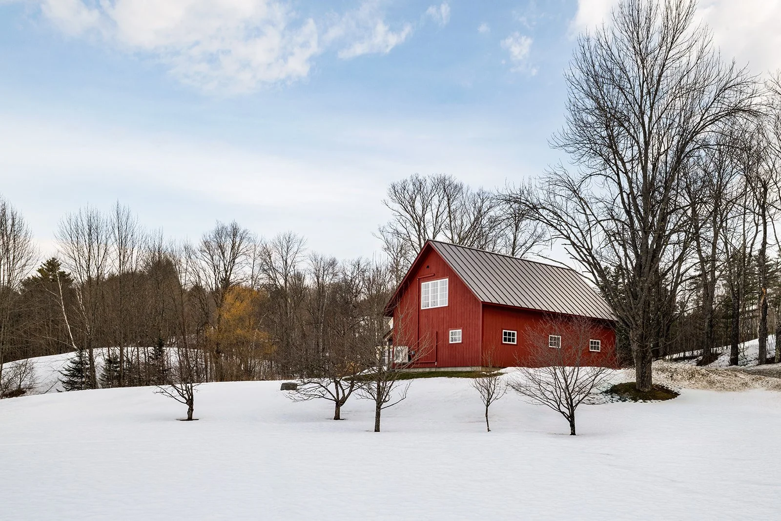 A red barn with white windows and a gray metal roof sits atop a small hill looking out onto a growing orchard and the Vermont hillside.