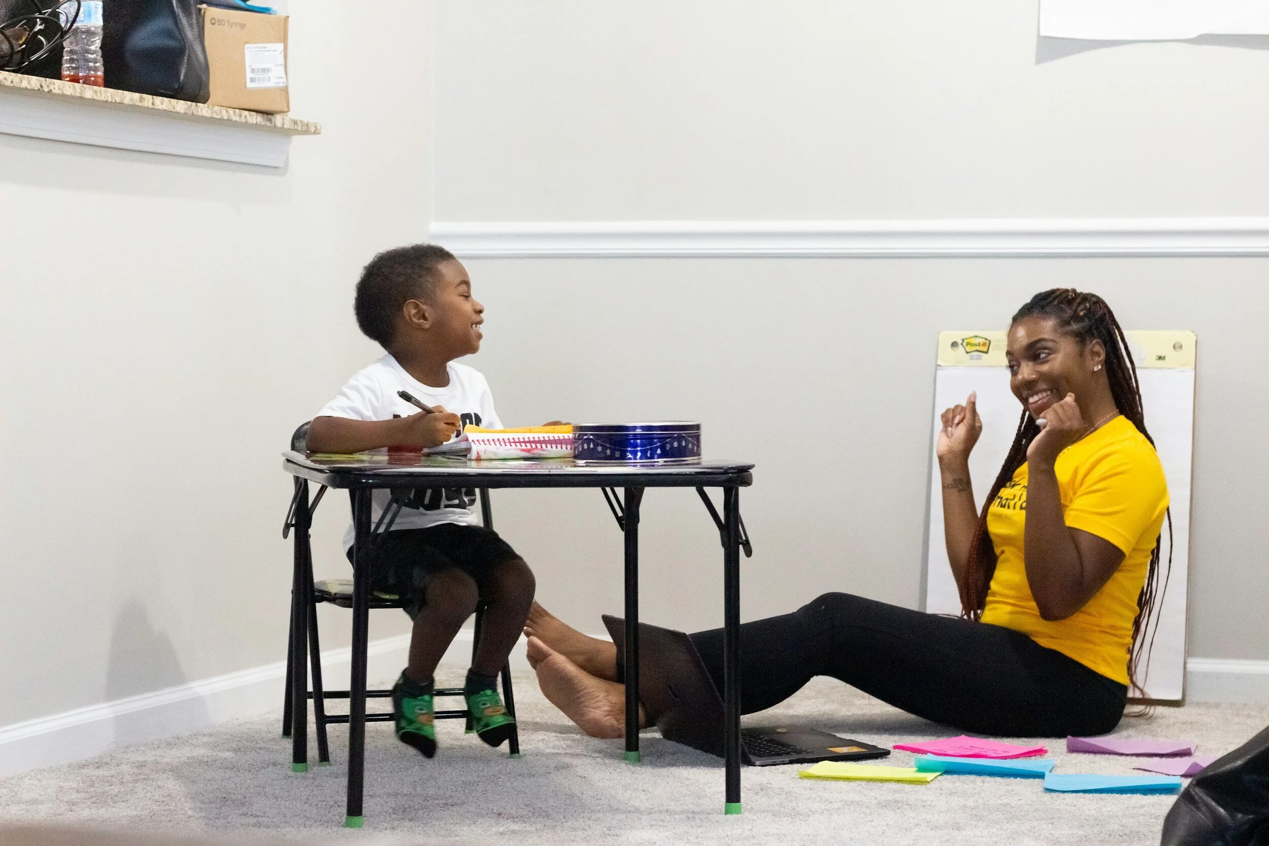 A child sitting at a small table with a notebook and a tin can, smiling at an adult sitting on the floor, who is smiling back. The adult wears a yellow shirt and has a laptop and colorful papers around them. The setting appears to be a home or classroom.