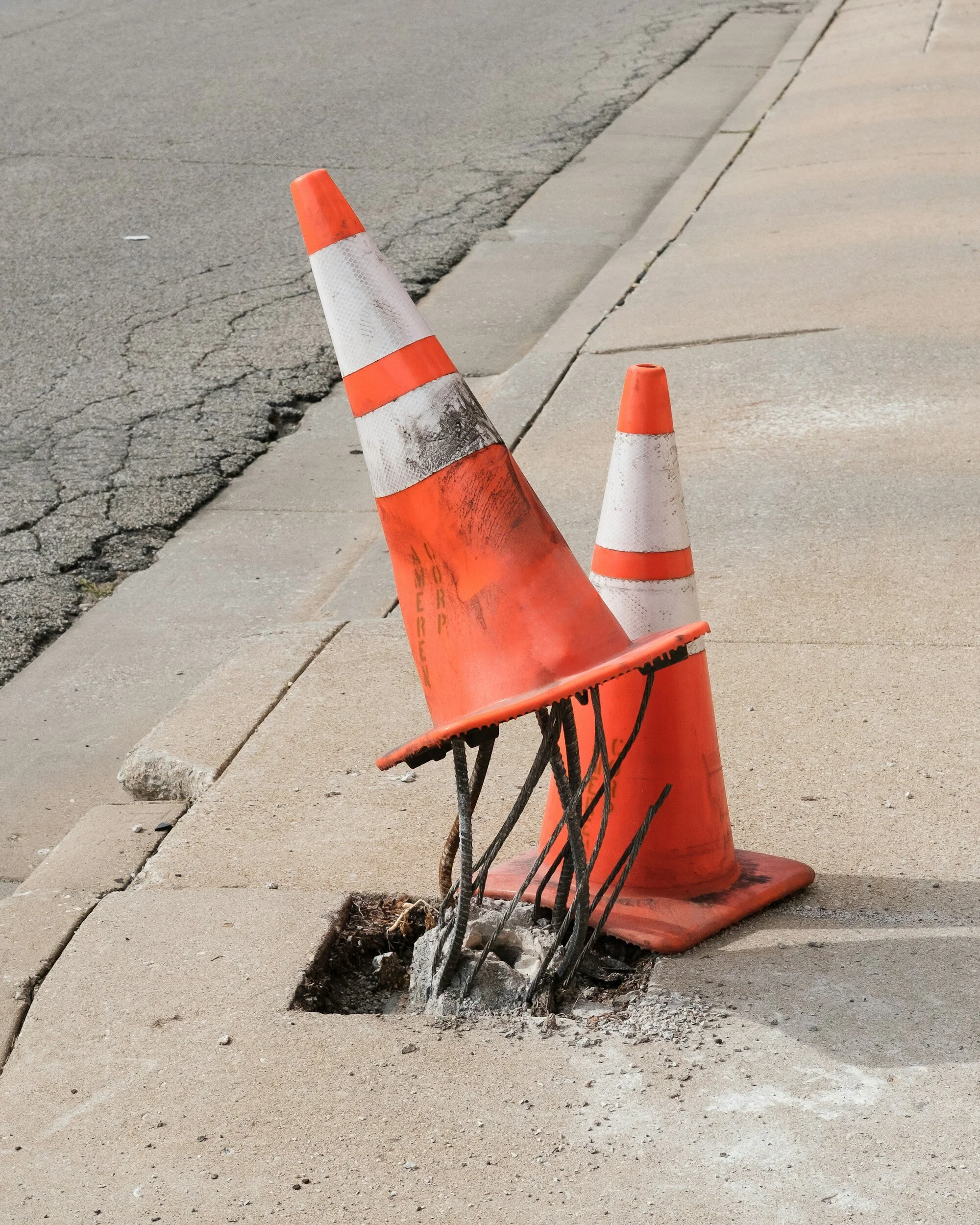 Two orange traffic cones on a sidewalk; one is covering exposed metal rebar protruding from a hole.