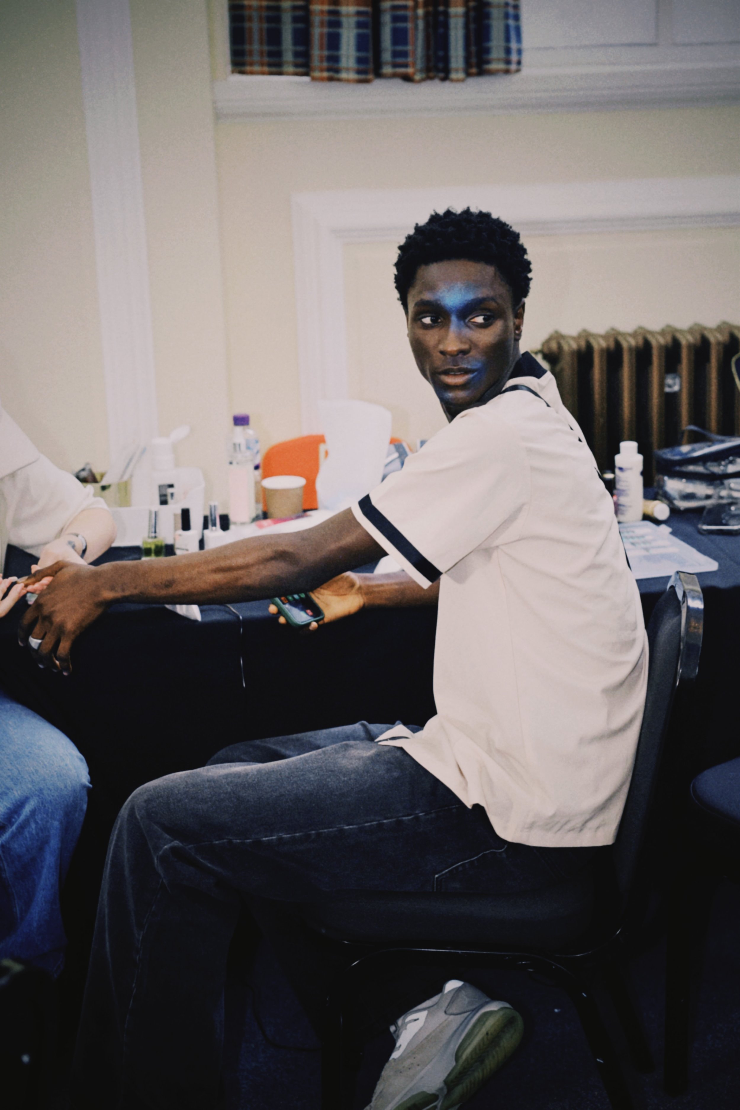 A young man with dark skin and curly black hair seated in a room with makeup and skincare products on the table behind him, wearing a white shirt with black trim and gray jeans, holding a smartphone, and looking to his right.