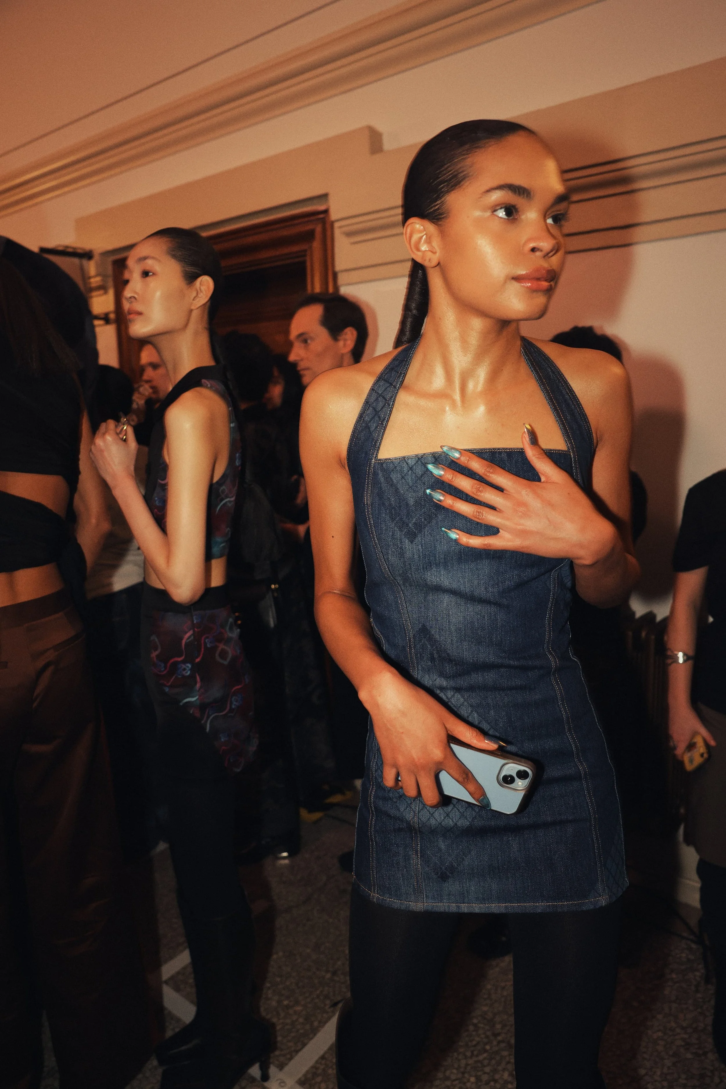 A woman in a denim dress holding a smartphone, with other people in a crowded indoor setting.