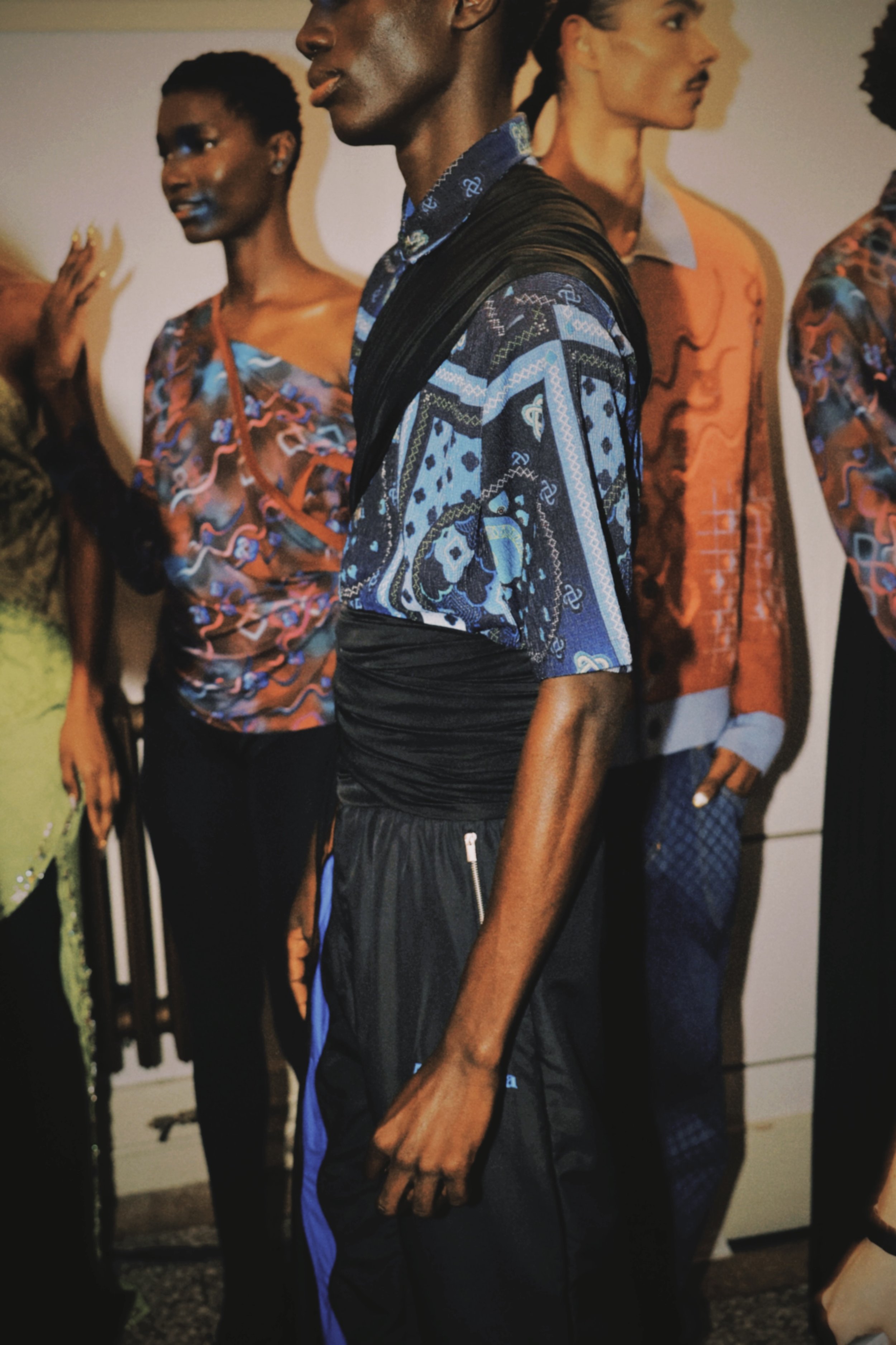 A man dressed in a vibrant, patterned shirt and black athletic pants standing among mannequins dressed in colorful, patterned clothing, in what appears to be a display or exhibit.