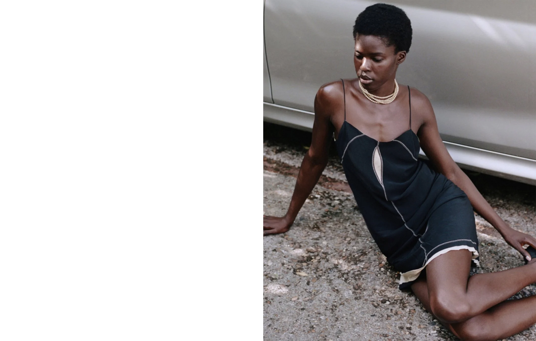 A woman with short curly black hair wearing a navy blue slip dress with white piping, kneeling on gravel ground beside a gray car, looking down with a serious expression.