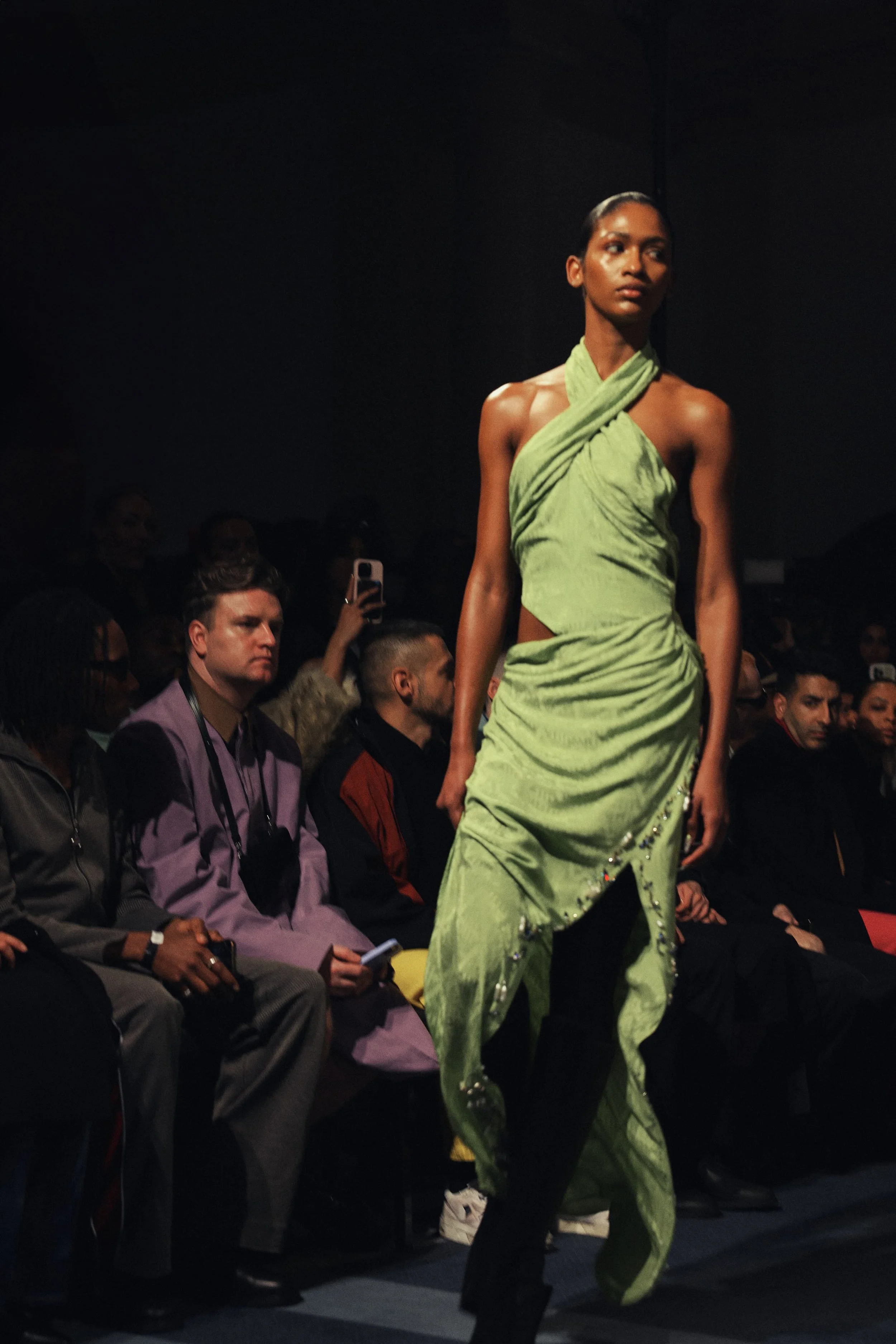 A fashion model in a lime green dress walks down the runway at a fashion show, with audience members sitting and watching.