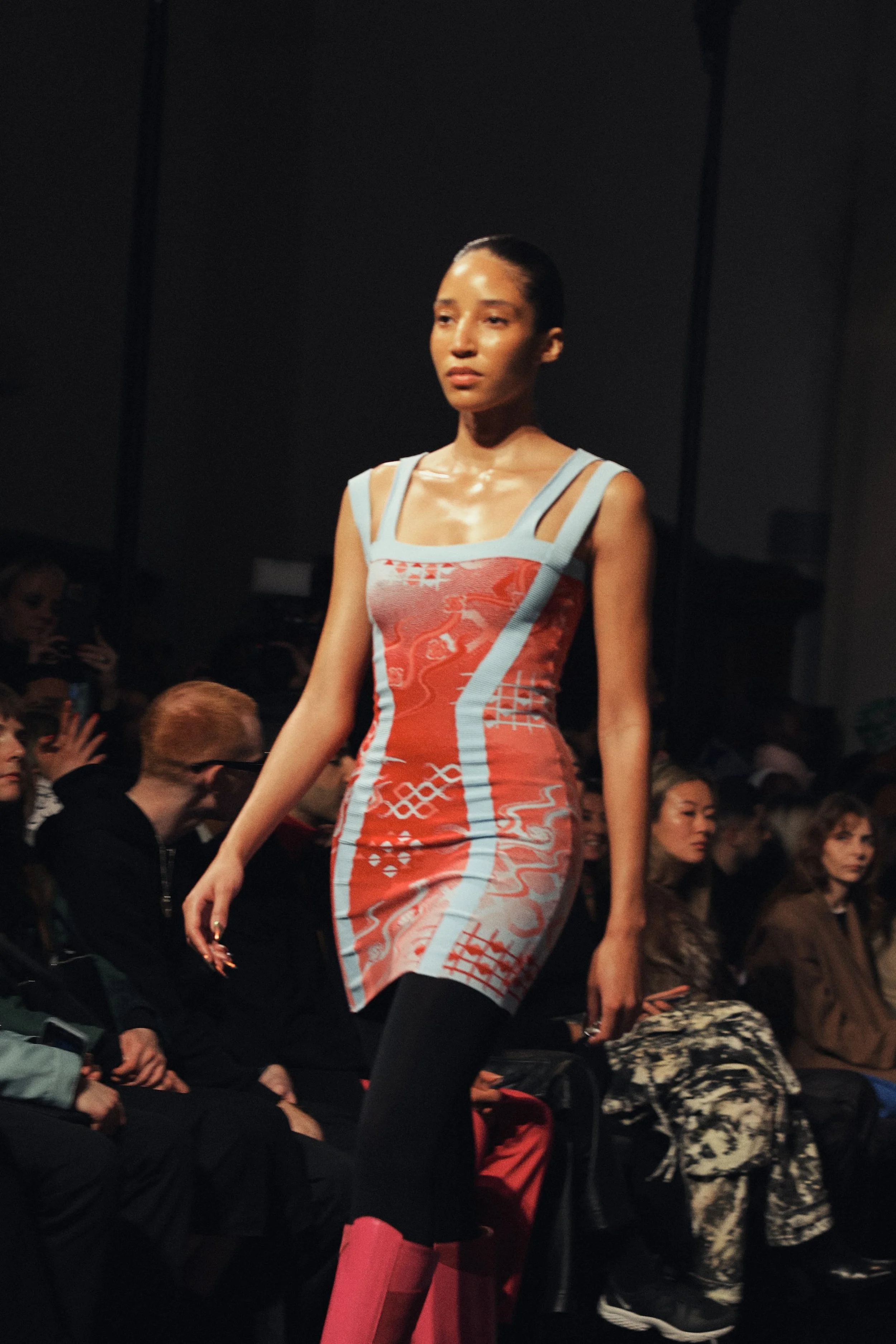 A fashion model walking on runway at a fashion show, wearing a colorful, patterned dress with black leggings and pink boots, with audience in background.