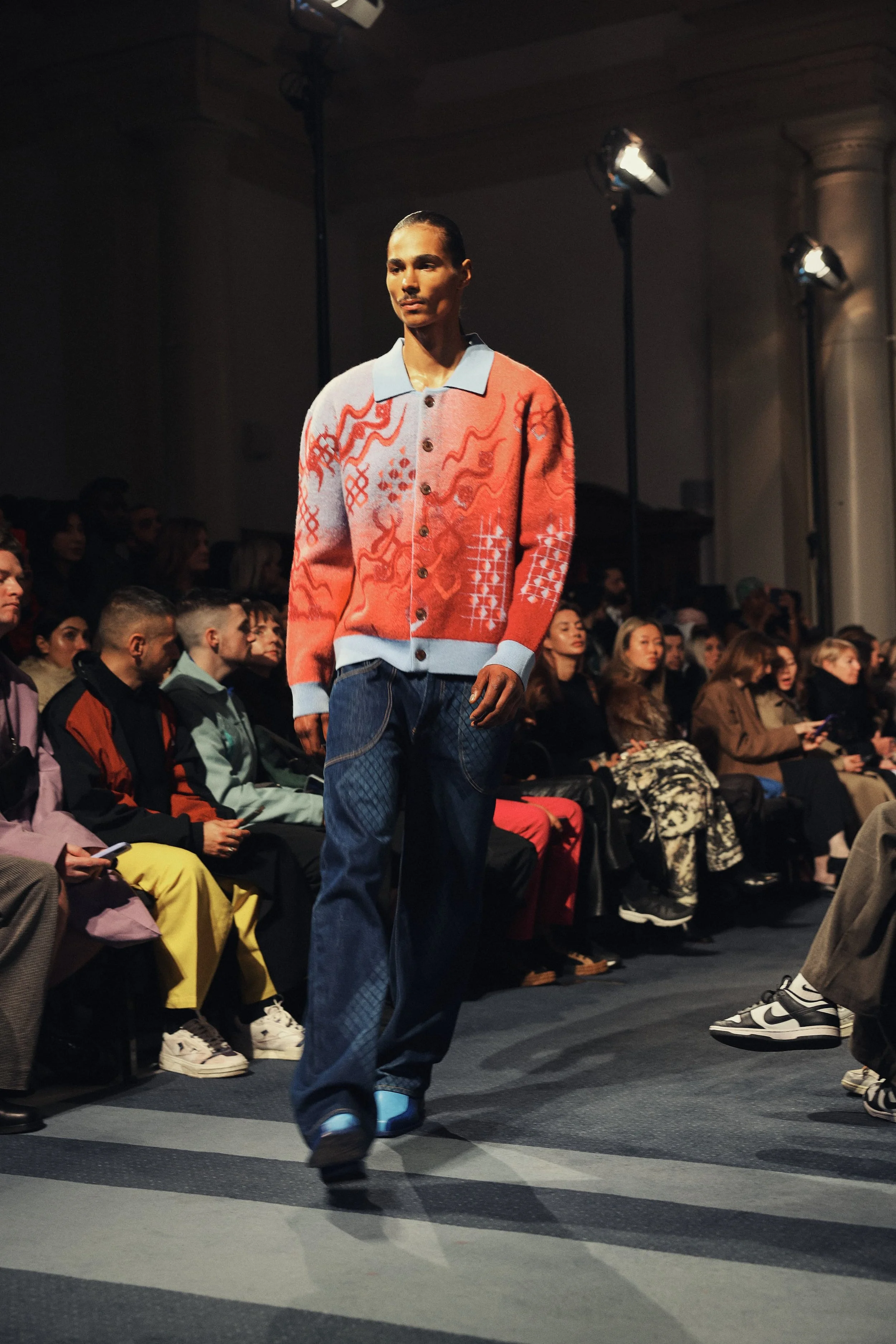 Model walking down runway during a fashion show, wearing a colorful patterned cardigan with a light blue collar and cuffs, wide-leg jeans, and sneakers, with audience seated on both sides.