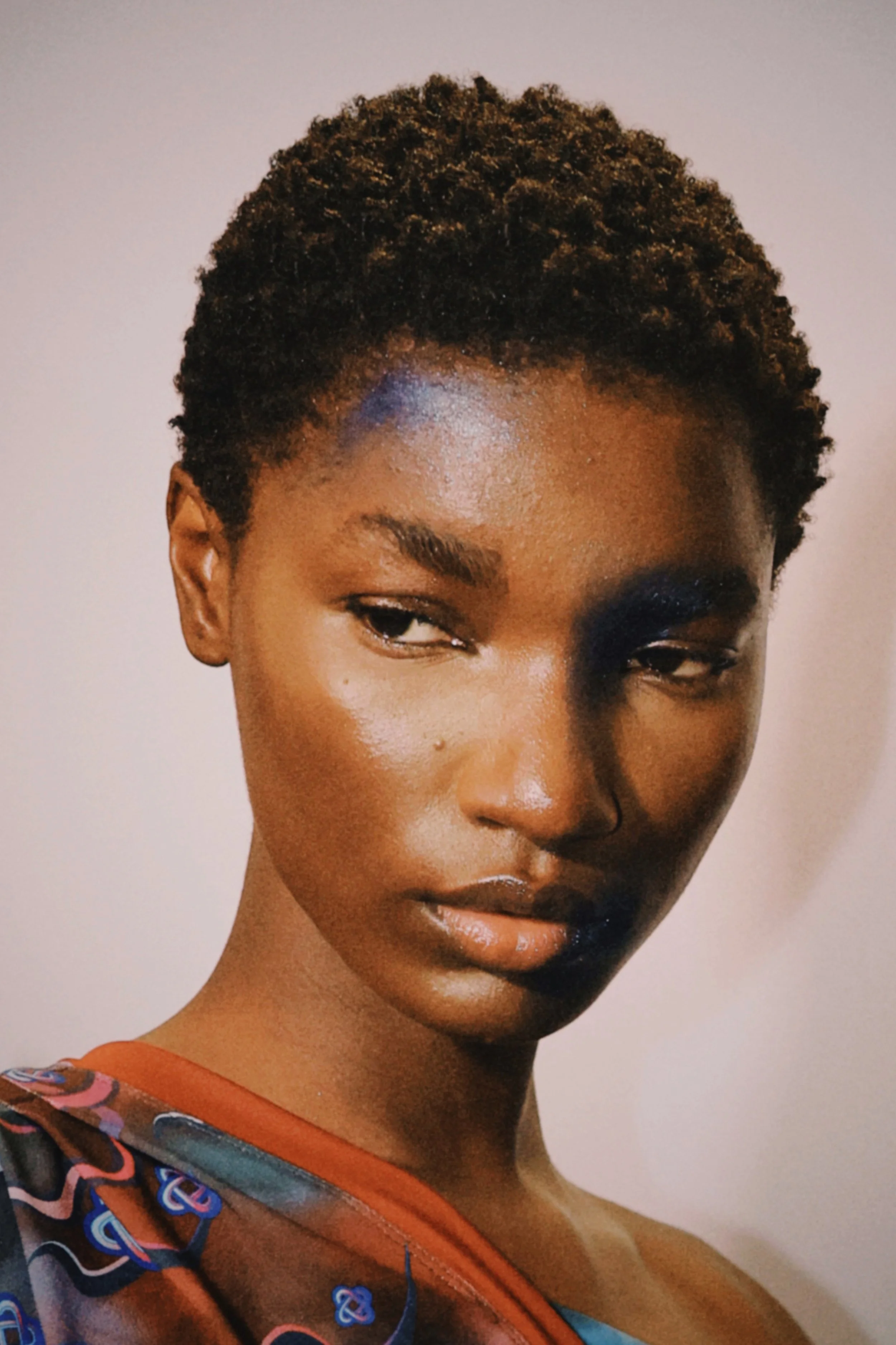 Close-up portrait of a young woman with short, curly hair, looking slightly to the side with a serious expression, wearing a colorful patterned top.