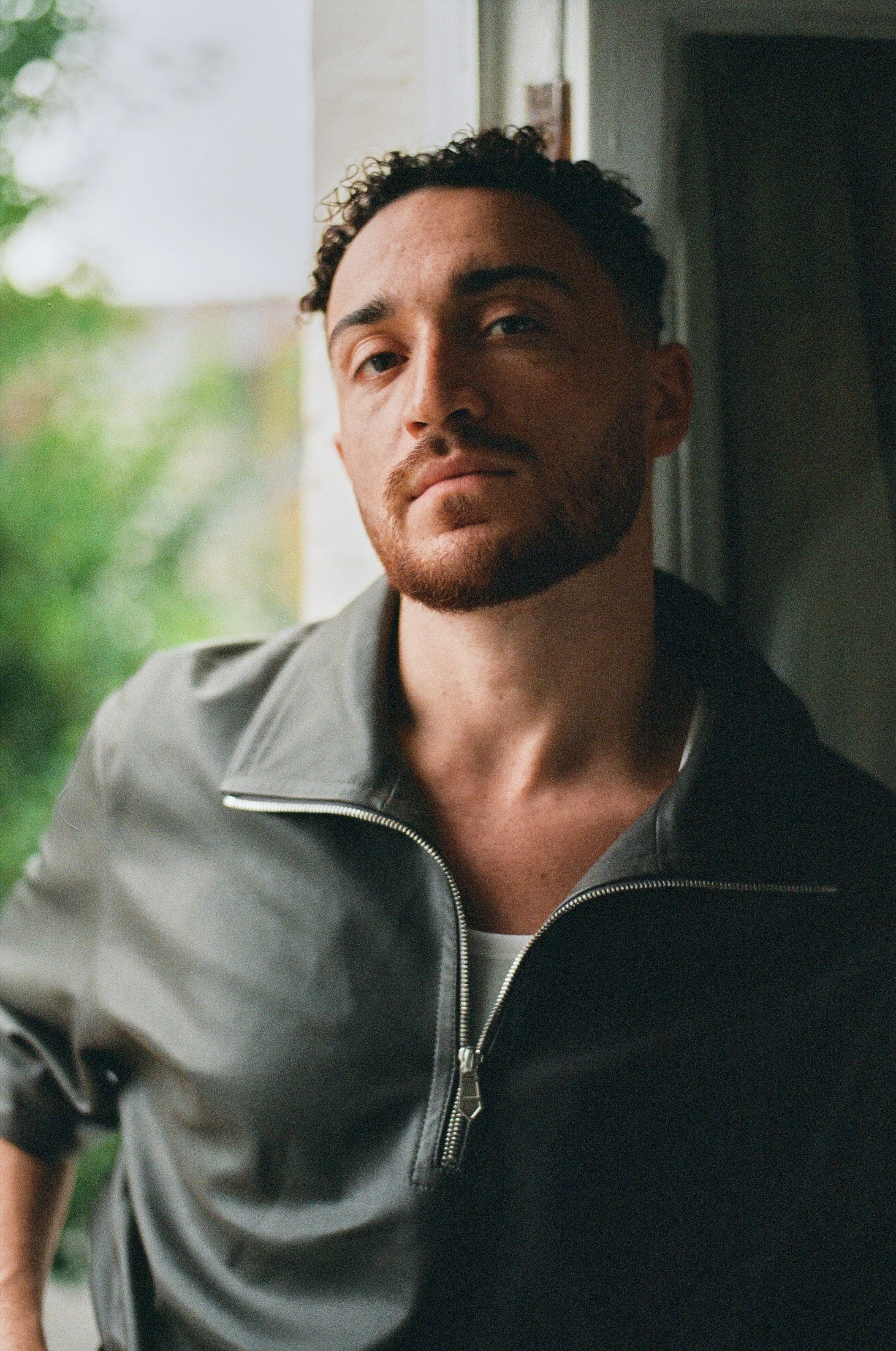 A man with short curly hair and a beard, looking relaxed, wearing a gray zip-up jacket, sitting by a window with greenery outside.