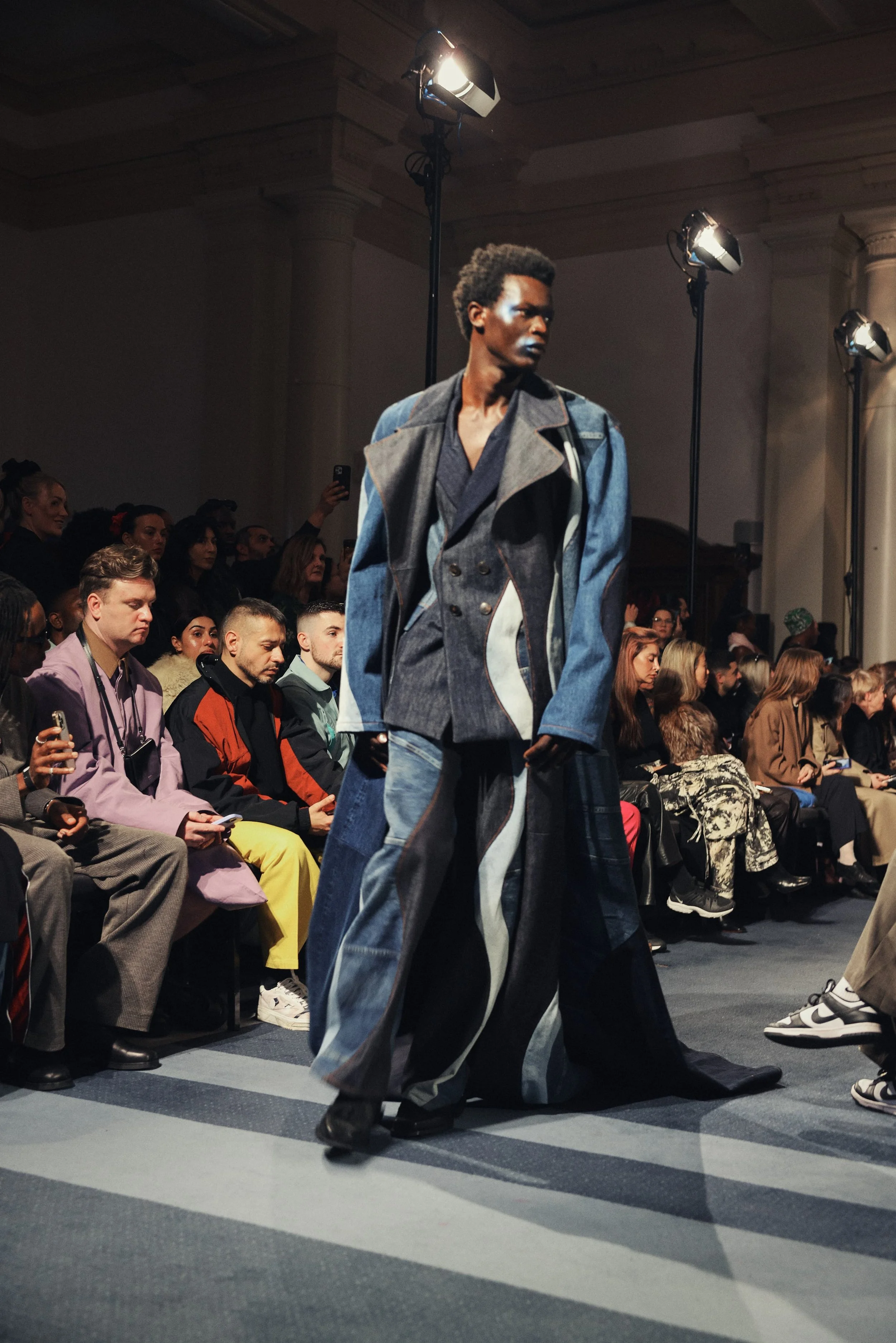 A male runway model walking at a fashion show, wearing a layered denim and gray coat with wide-legged jeans, on a runway with an audience watching.