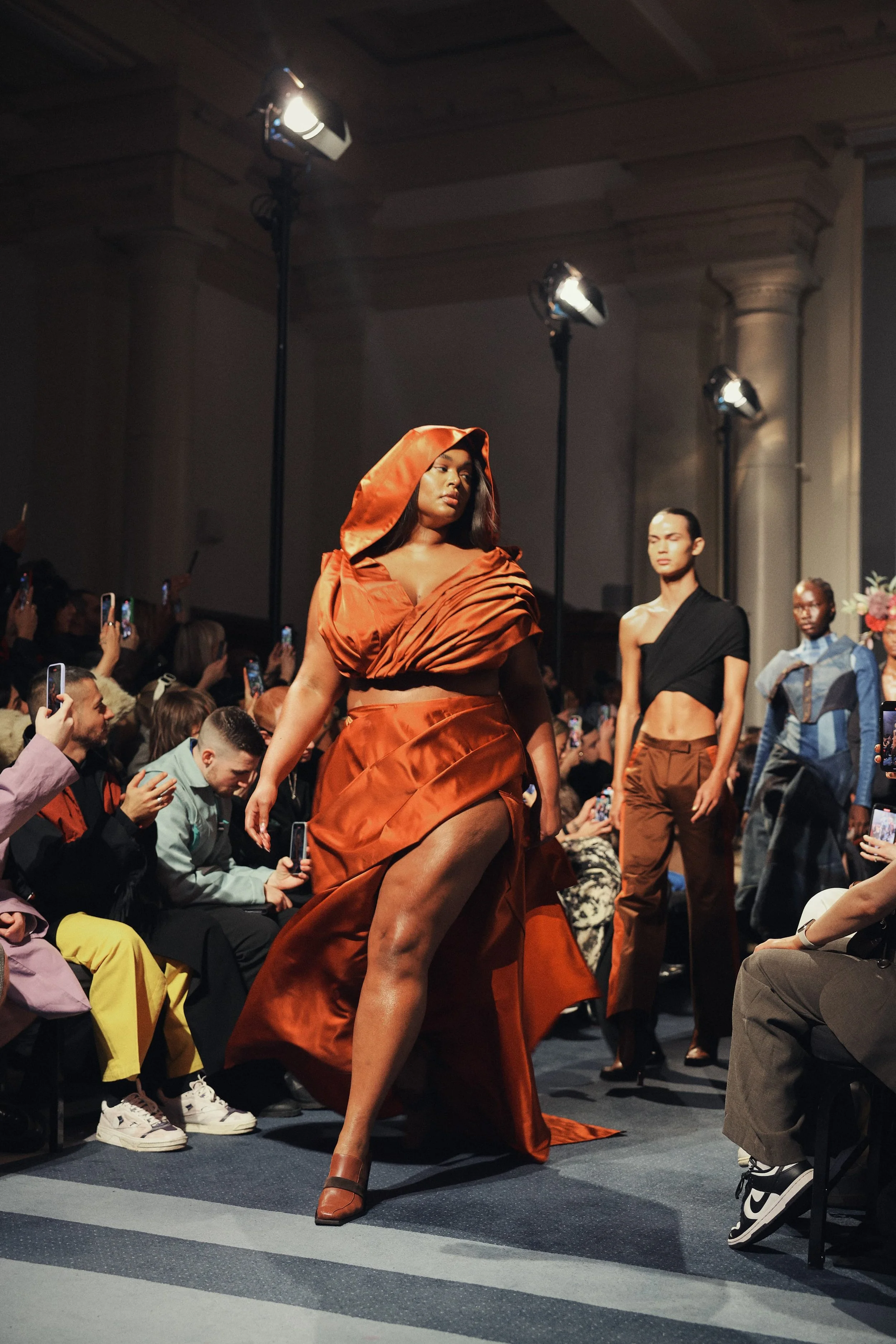 A diverse group of models participating in a fashion runway show, with an audience taking photos. The models are wearing high fashion clothing, with the central model in an elaborate orange outfit with a hood and thigh-high slit.