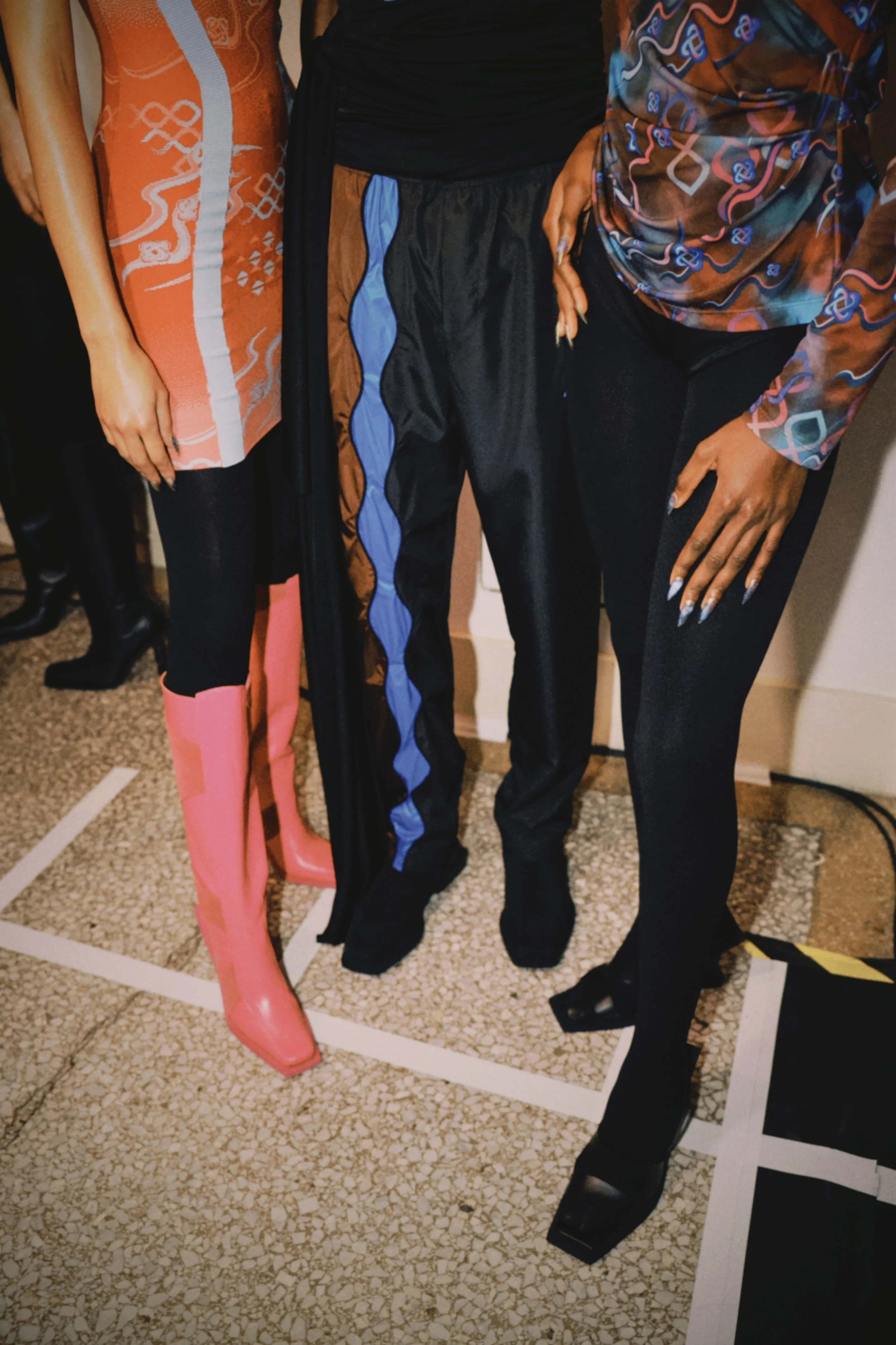 Close-up of three women standing together, focusing on their colorful and fashionable outfits, including pink boots, blue and black pants, and patterned tops.