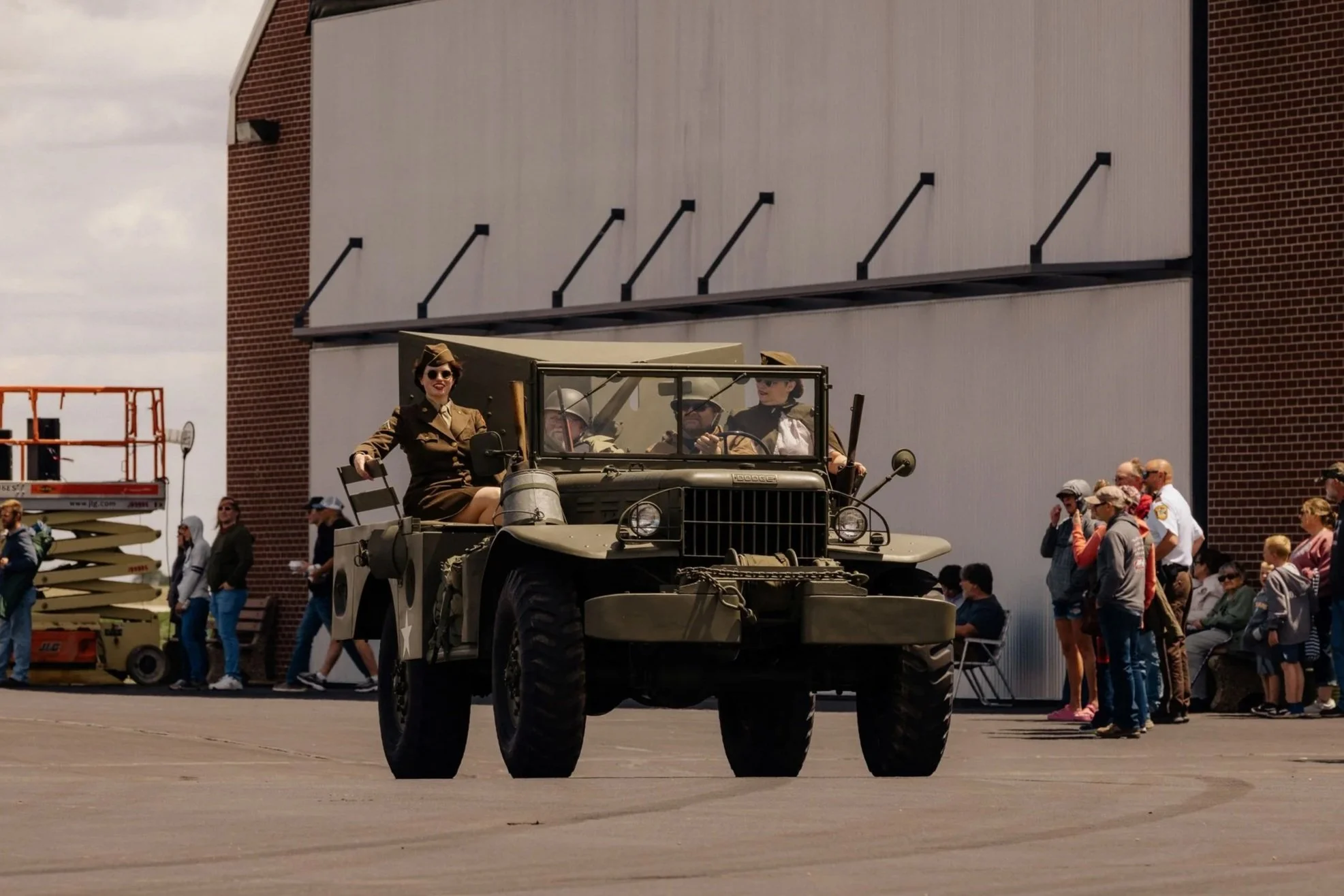 A vintage military-style jeep with four women dressed as soldiers inside, driving down a street during a parade or event. There are people watching on the side of the road, some taking photos, with a brick building and a large blank wall in the background.