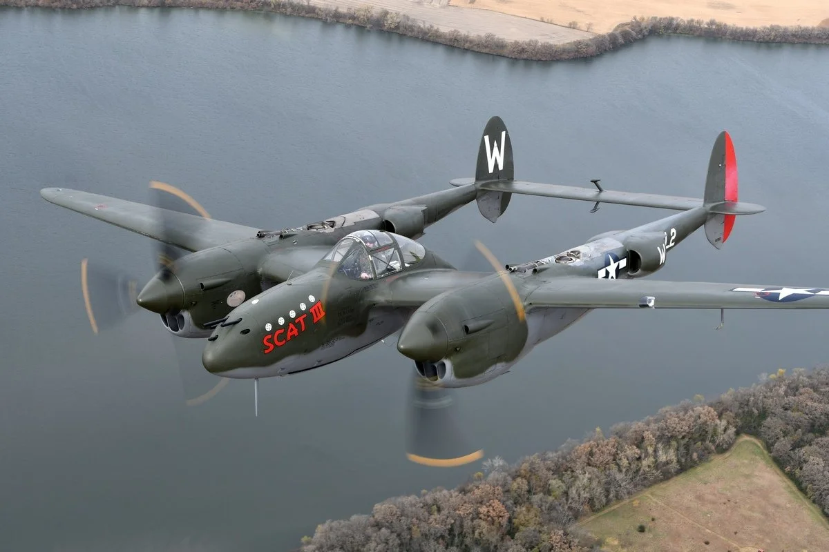 A two-engine P-38 Lightning military aircraft flying over a body of water, with a landscape of trees and fields below.