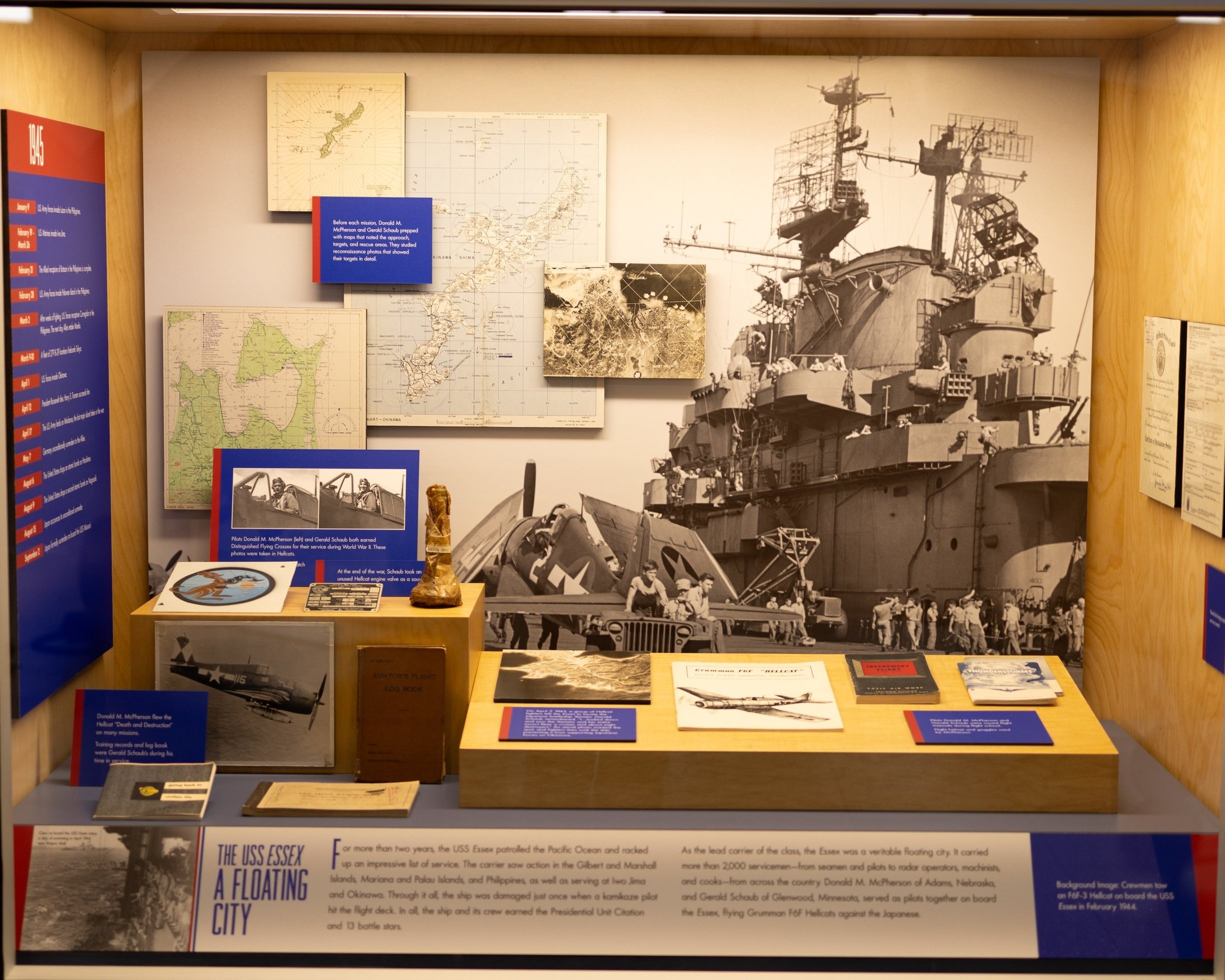 Historical display about the USS Essex, a World War II aircraft carrier, including maps, photographs, documents, model aircraft, and informational text about its service and crew, with a large black-and-white photo of the ship in a navy museum exhibit.