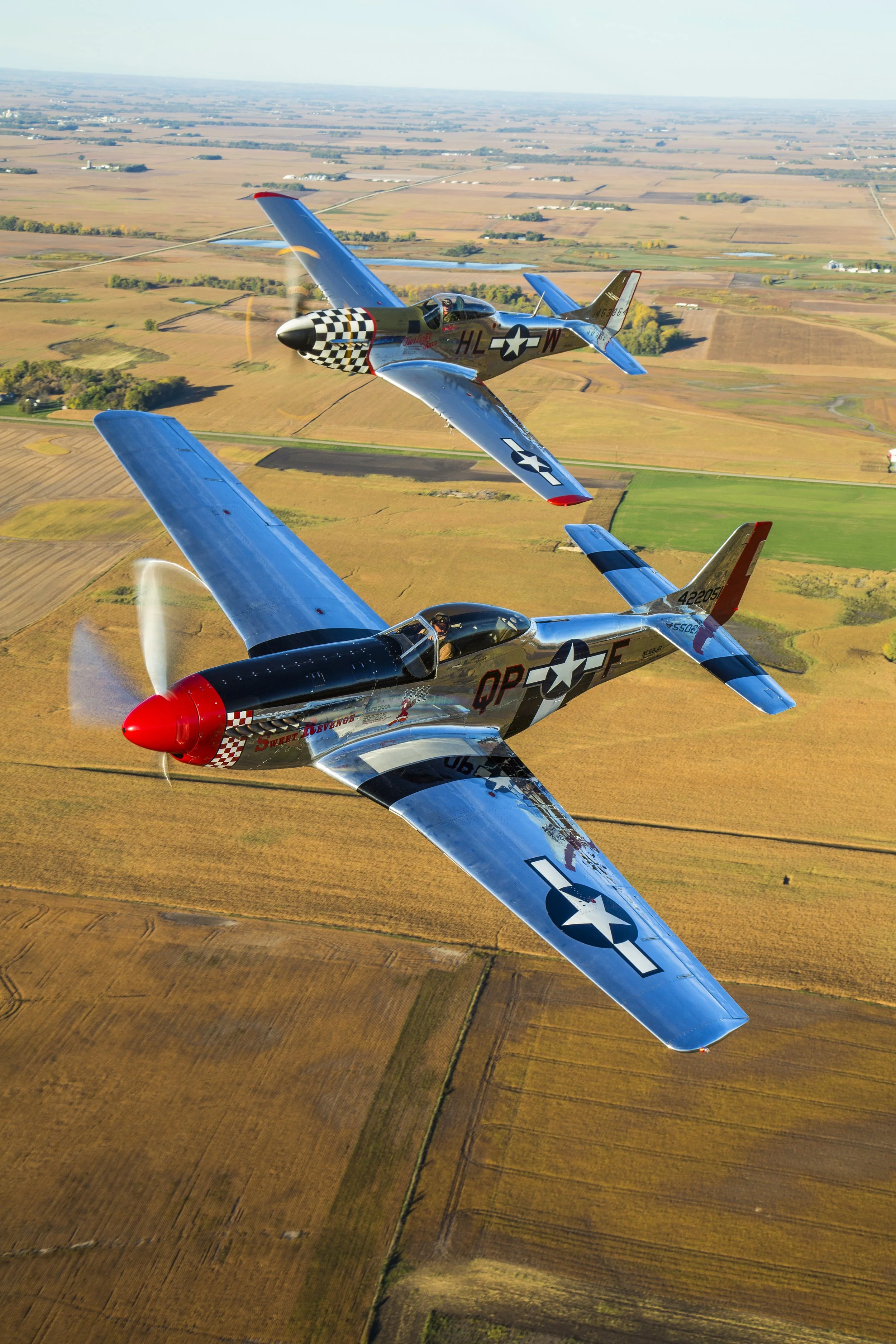 Two P-51 Mustangs flying in formation over a rural landscape with fields and farmland.