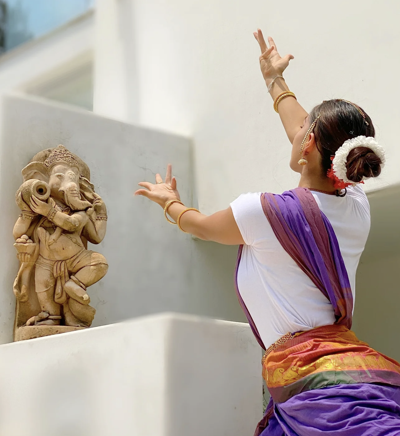 Mujer bailando frente a una estatua de un dios hindú, vestida con ropa tradicional, adornada con flores en el cabello y joyas.