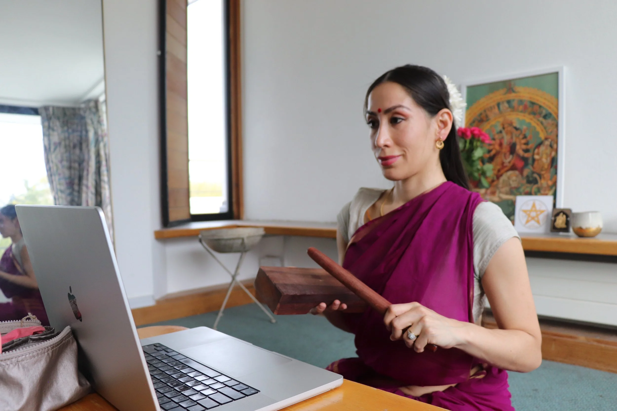 Mujer en traje tradicional indio usando una laptop, sosteniendo dos bloques de madera similar a un instrumento. Detrás hay una pintura religiosa hindú y flores en una mesa.