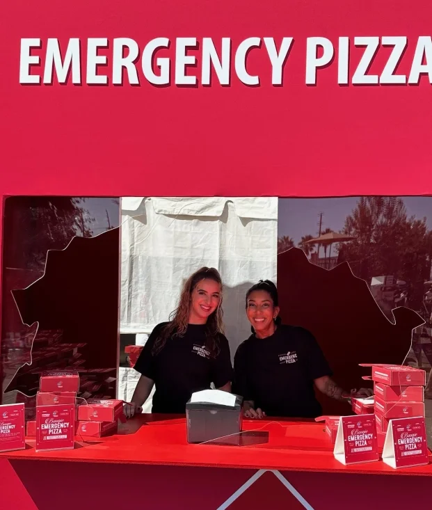 Two people stand at a red "Emergency Pizza" booth with a Domino's logo, distributing promotional boxes.