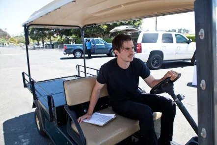 A man sitting on a golf cart with a clipboard beside him, holding the steering wheel, in a parking lot with cars and trees in the background.