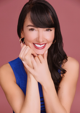 Woman with dark hair smiling, wearing a blue dress, and posing with her hands near her face against a pink background.