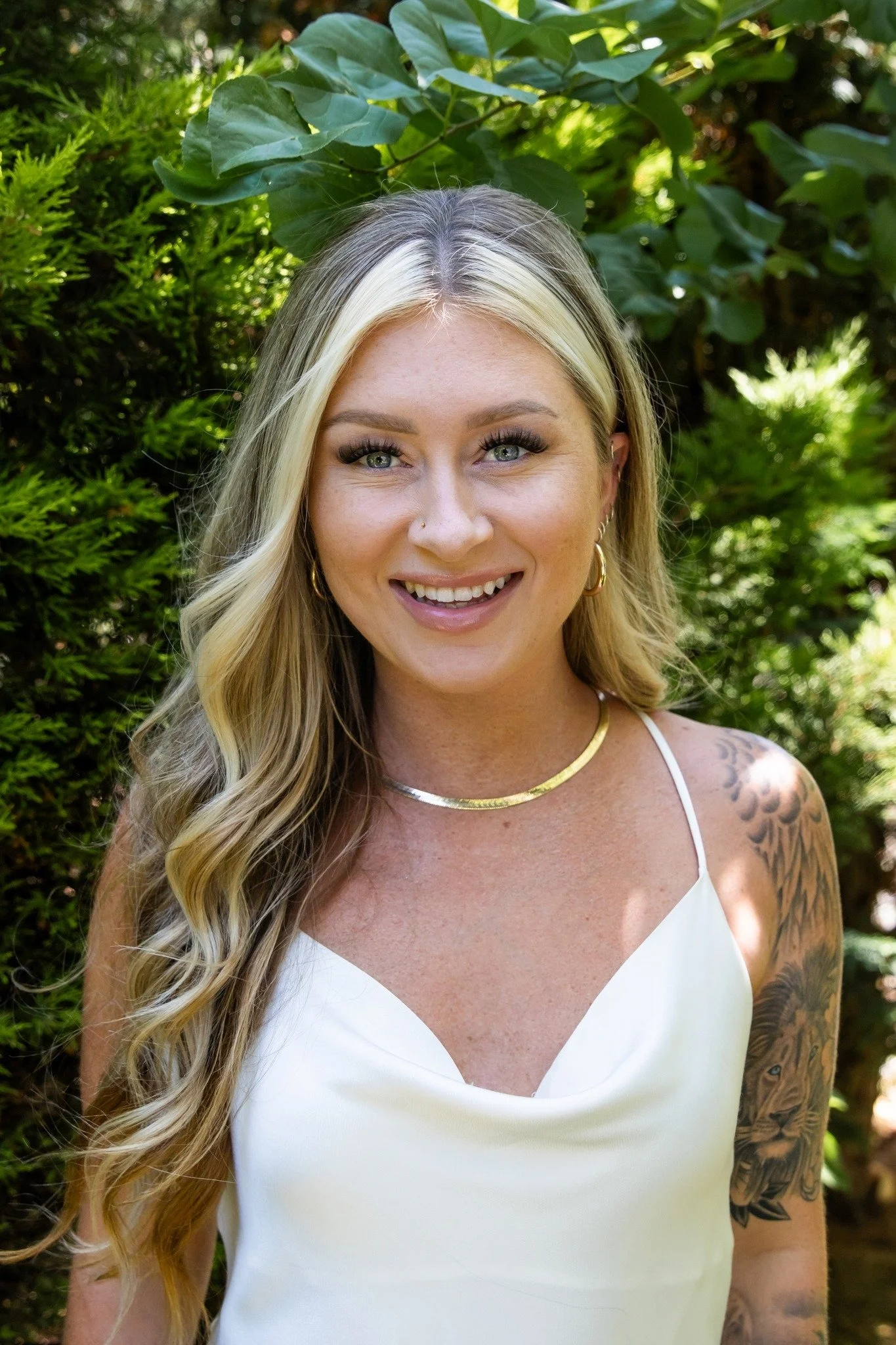 A smiling blonde woman with wavy hair, wearing a white spaghetti strap top, gold jewelry, and a tattoo on her arm, stands outdoors in front of green foliage.