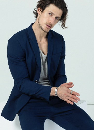 Man with brown hair wearing a navy blue suit and gray shirt, sitting and clasping hands with silver rings and bracelet.