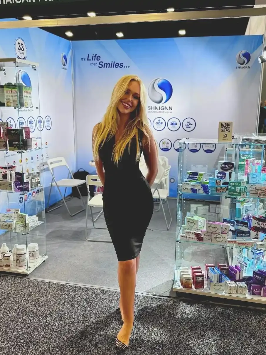 A woman standing in front of a booth with pharmaceutical products and a large banner for Shaigan Healthcare, smiling and dressed in a black dress.