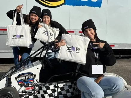 Three people posing with white zip tote bags beside a mini race car labeled "NASCAR Youth Series."