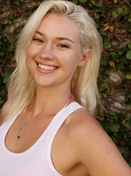 Person with blonde hair smiling outdoors, wearing a white tank top against a leafy background.