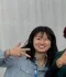 Smiling person with black hair making a peace sign with fingers, wearing a striped shirt and lanyard, standing indoors.