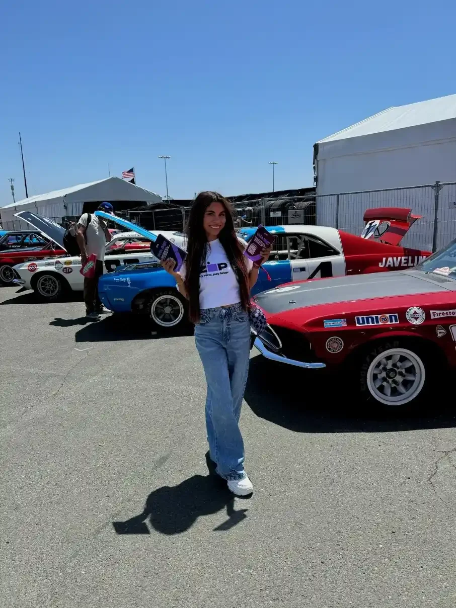 Woman holding pamphlets stands near classic cars at an outdoor car event under a clear blue sky.