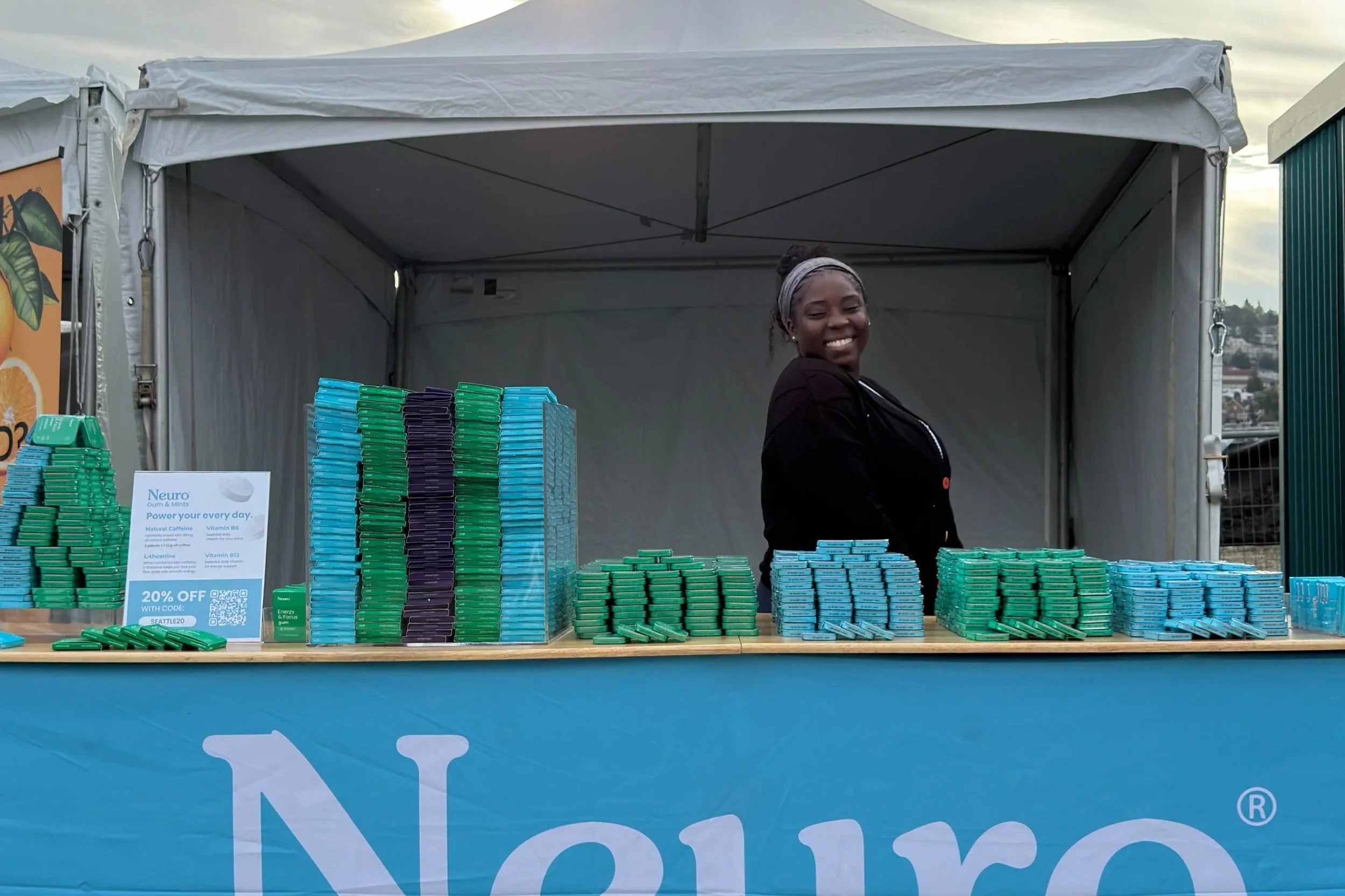 Woman smiling behind a booth selling gum and mints, with stacks of blue, green, and purple containers on the table, under a white tent at an outdoor event.