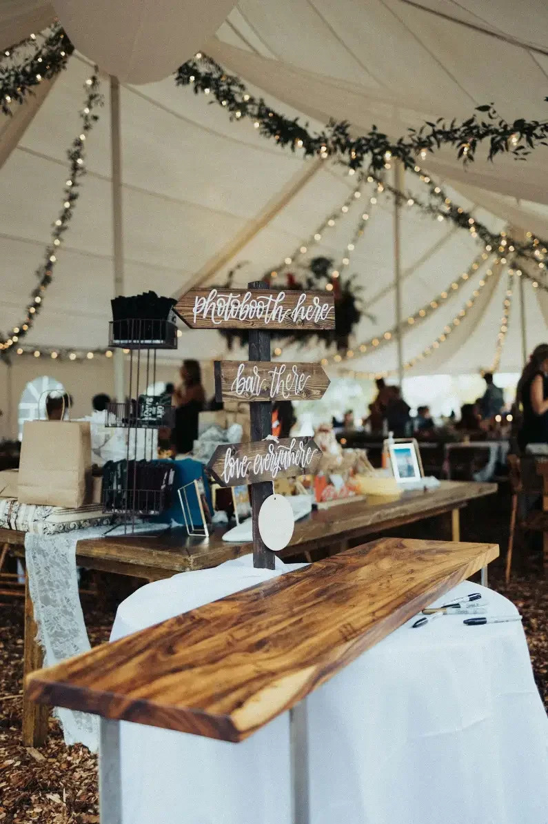 Indoor wedding reception with rustic decor, wooden directional sign reading "Photobooth here, bar there, love everywhere," wooden table, string lights, and guests in background.