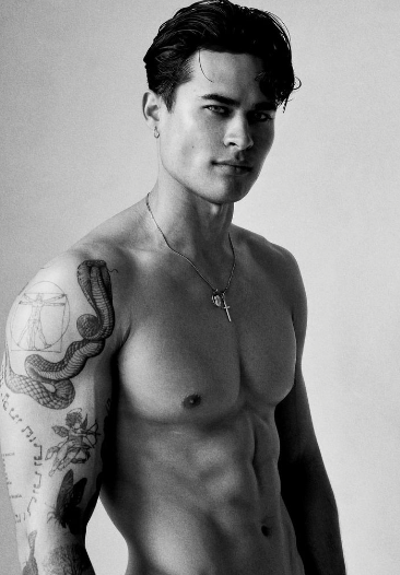 Black and white photo of a shirtless man with tattoos on his arm and chest, wearing a necklace.