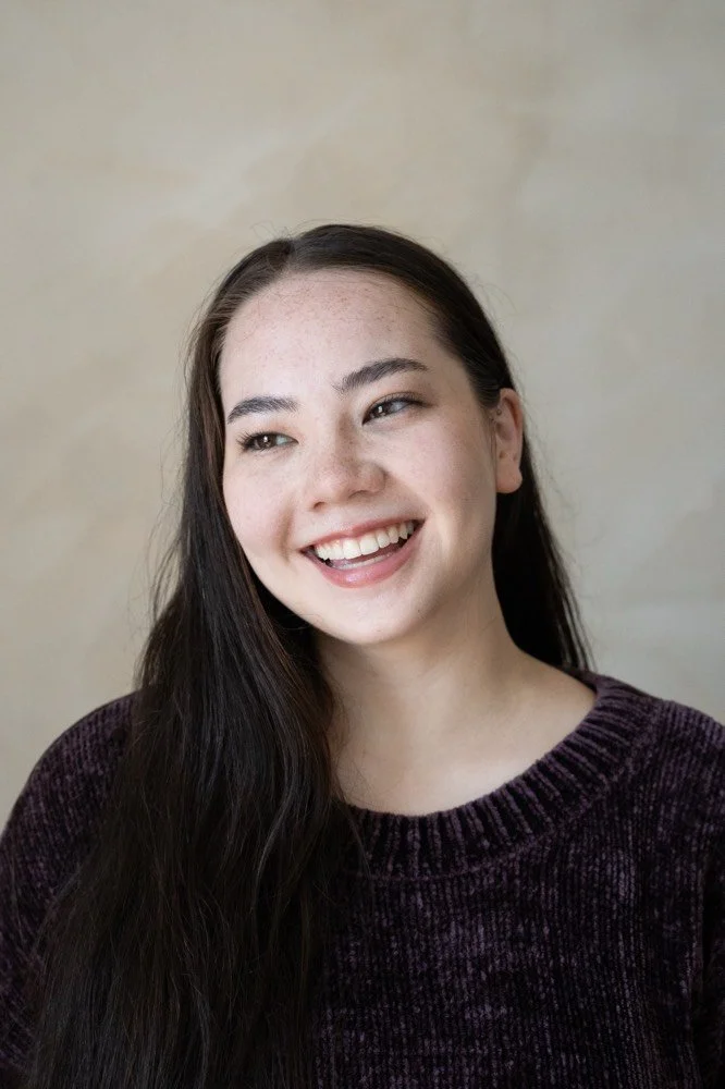 A young woman with long dark hair, smiling and looking to her left, wearing a dark purple sweater against a beige background.