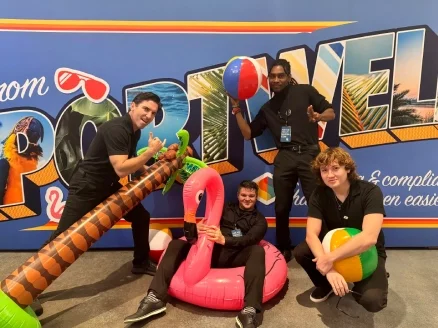 Four people posing with inflatable toys, including a palm tree, flamingo, and beach ball, in front of a colorful backdrop with tropical-themed lettering and images.