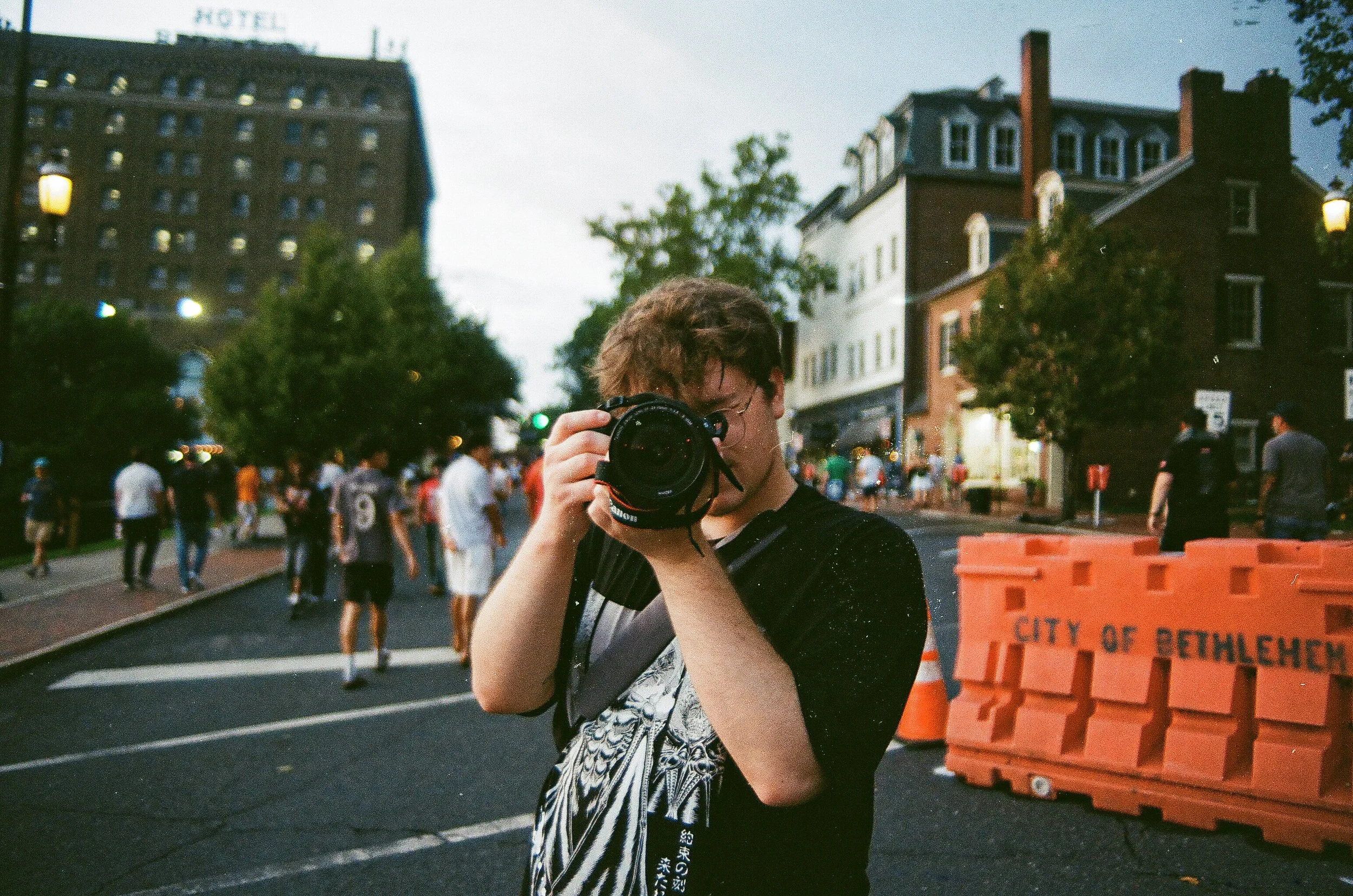 Person taking a photo with a camera on a city street during the evening, with shops, people, and orange barriers in the background.