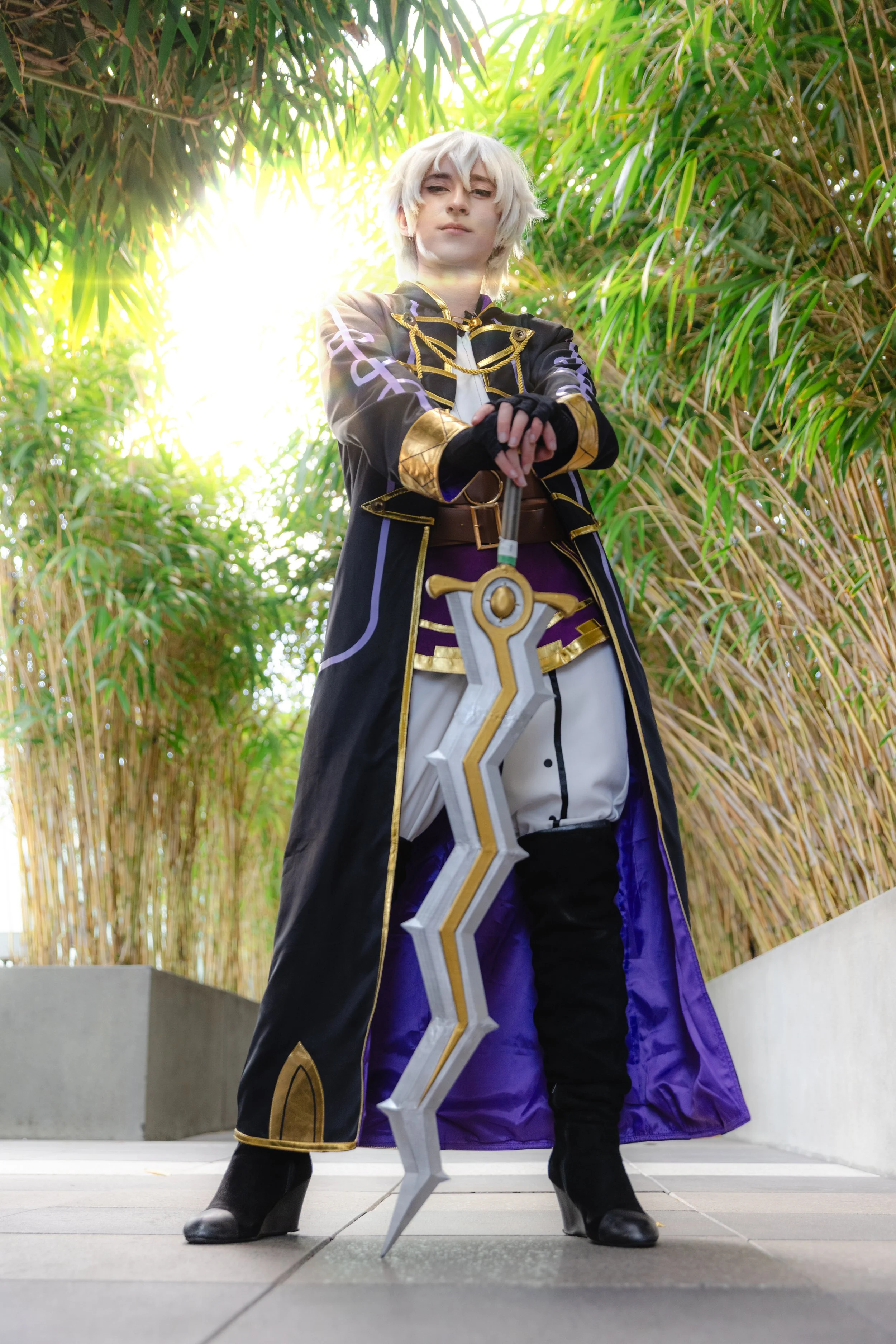 Person in cosplay costume holding a large sword, standing outdoors near bamboo plants, with sunlight behind them.