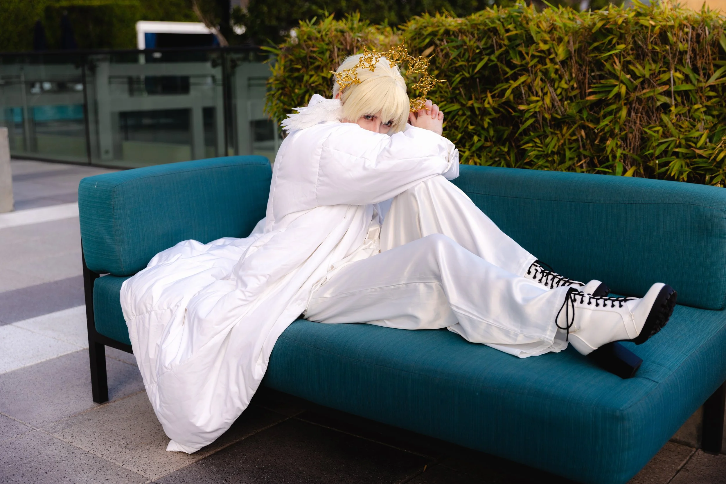 Person with blond hair wearing a golden crown, white angel costume, and black and white sneakers, sitting on a teal outdoor sofa with arms wrapped around their knees, looking at the camera surrounded by green shrubbery.