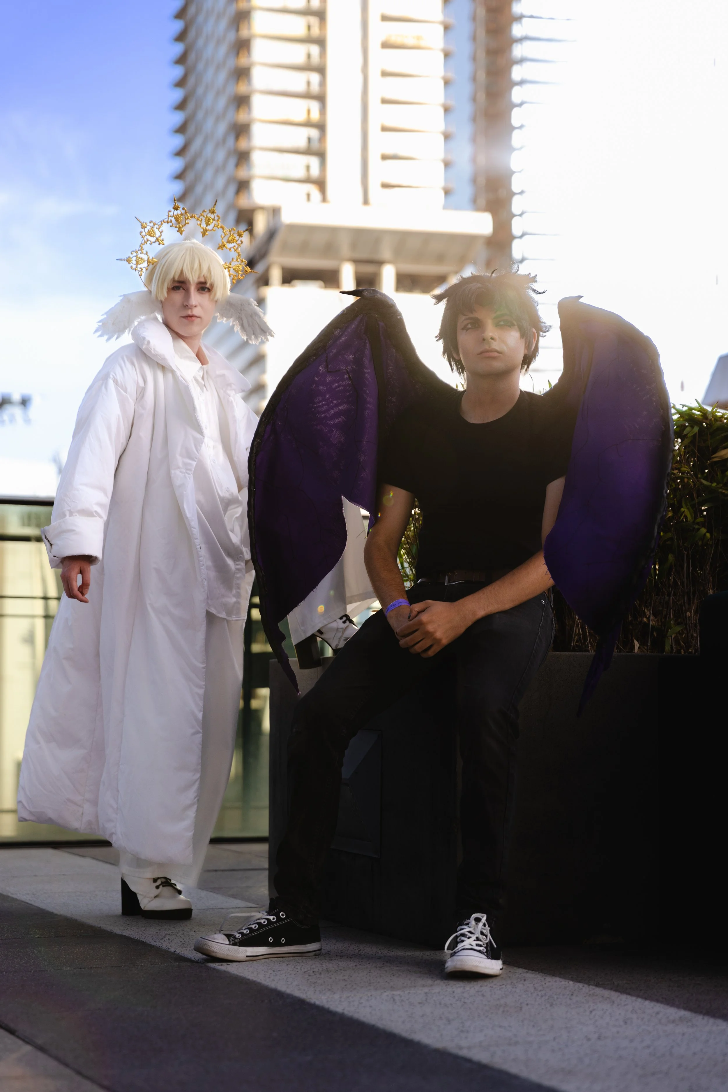 Two people in costume cosplay outdoors in front of a tall building, one dressed as an angel with a halo, white wings, and a white robe, and the other as a demon with purple wings and black clothing, both standing and sitting on a ledge.