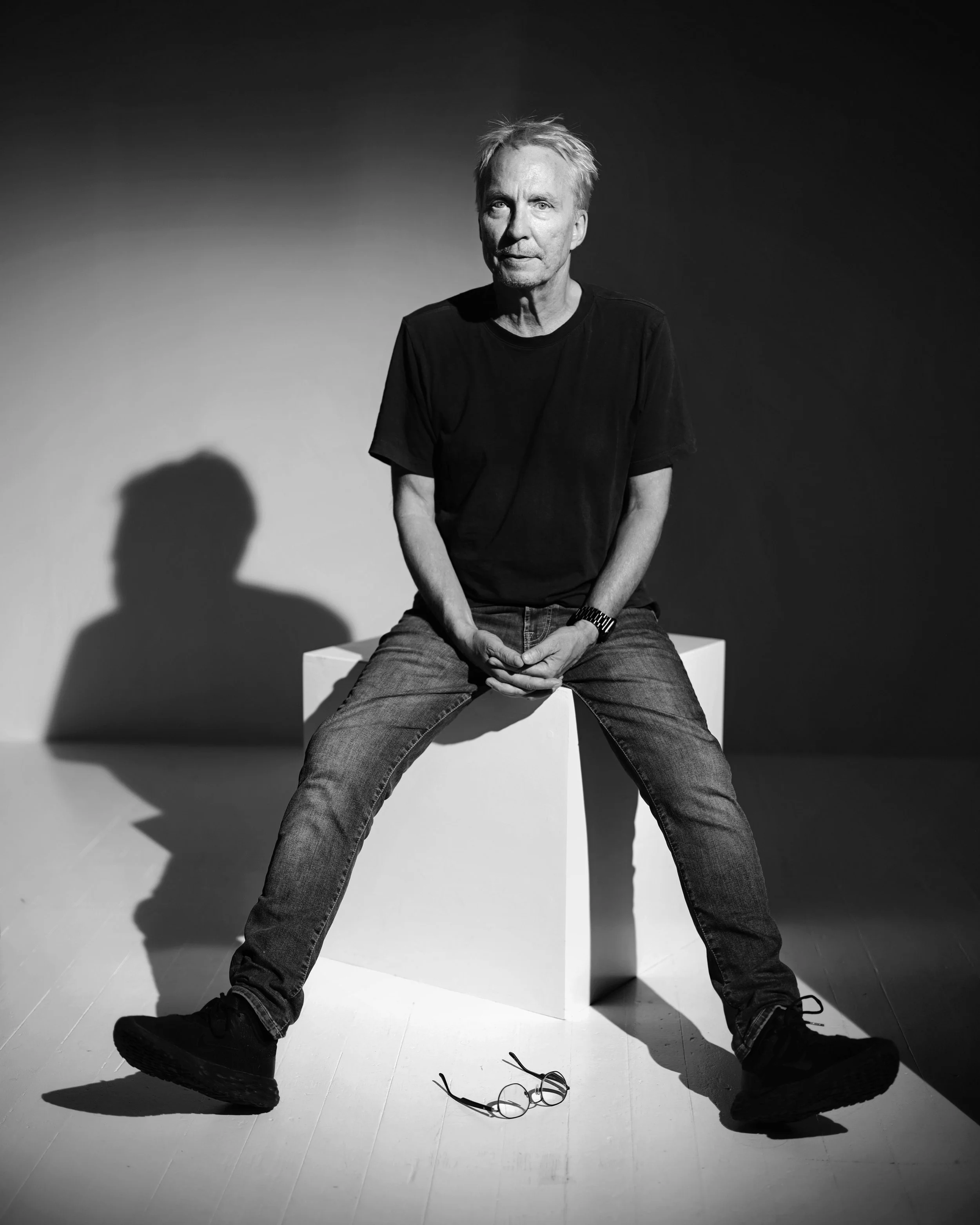 En svartvit bild av en äldre man sittande på en vit block, med glasögon på golvet framför honom. Mannen har ljus hår, bär en mörk T-shirt och jeans, och sitter med benen isär.