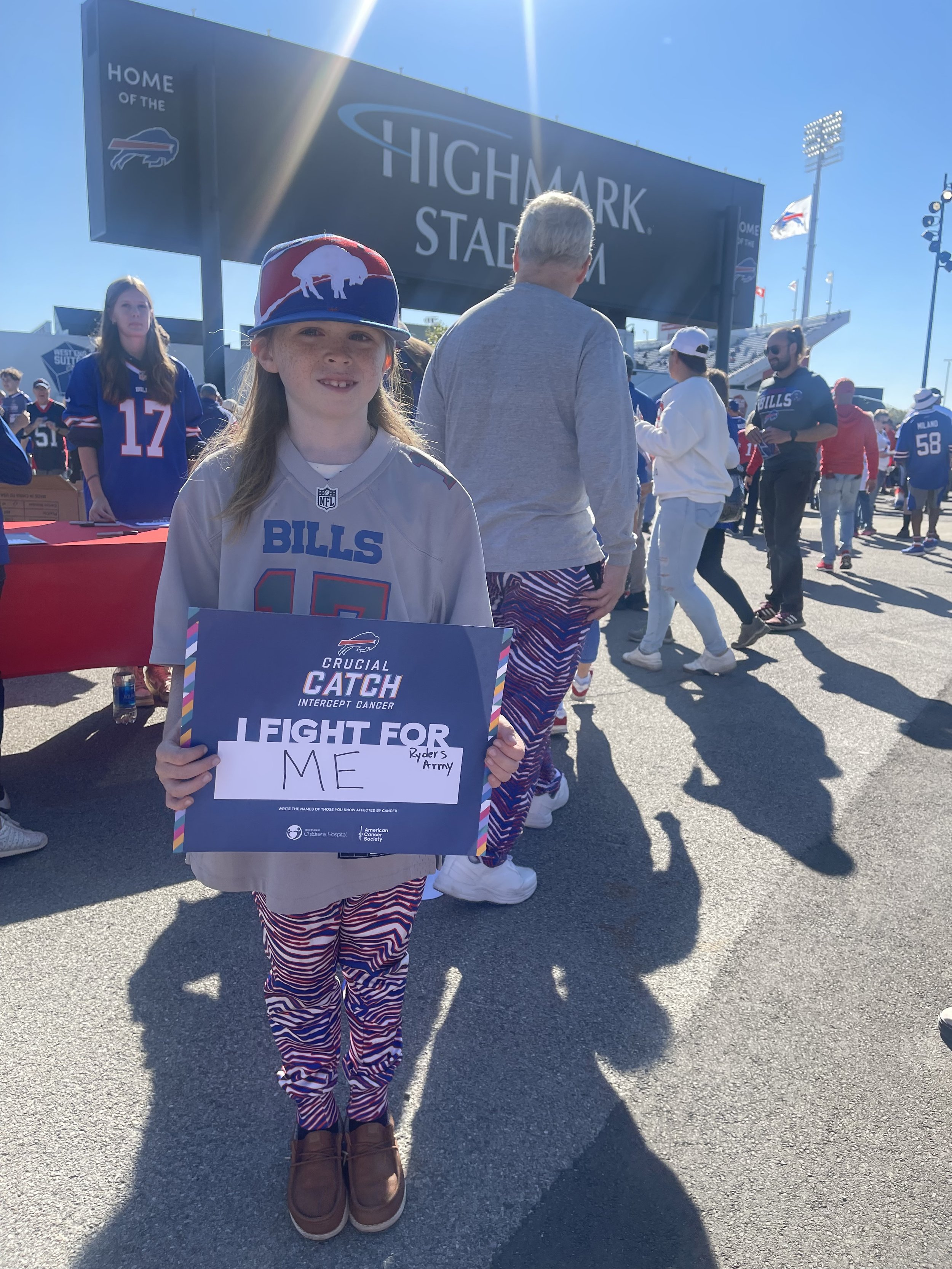 A child wearing a Buffalo Bills jersey and cap holds a sign for the "Crucial Catch Intercept Cancer" initiative, reading "I fight for me, Ryder's Army," outside Highmark Stadium.