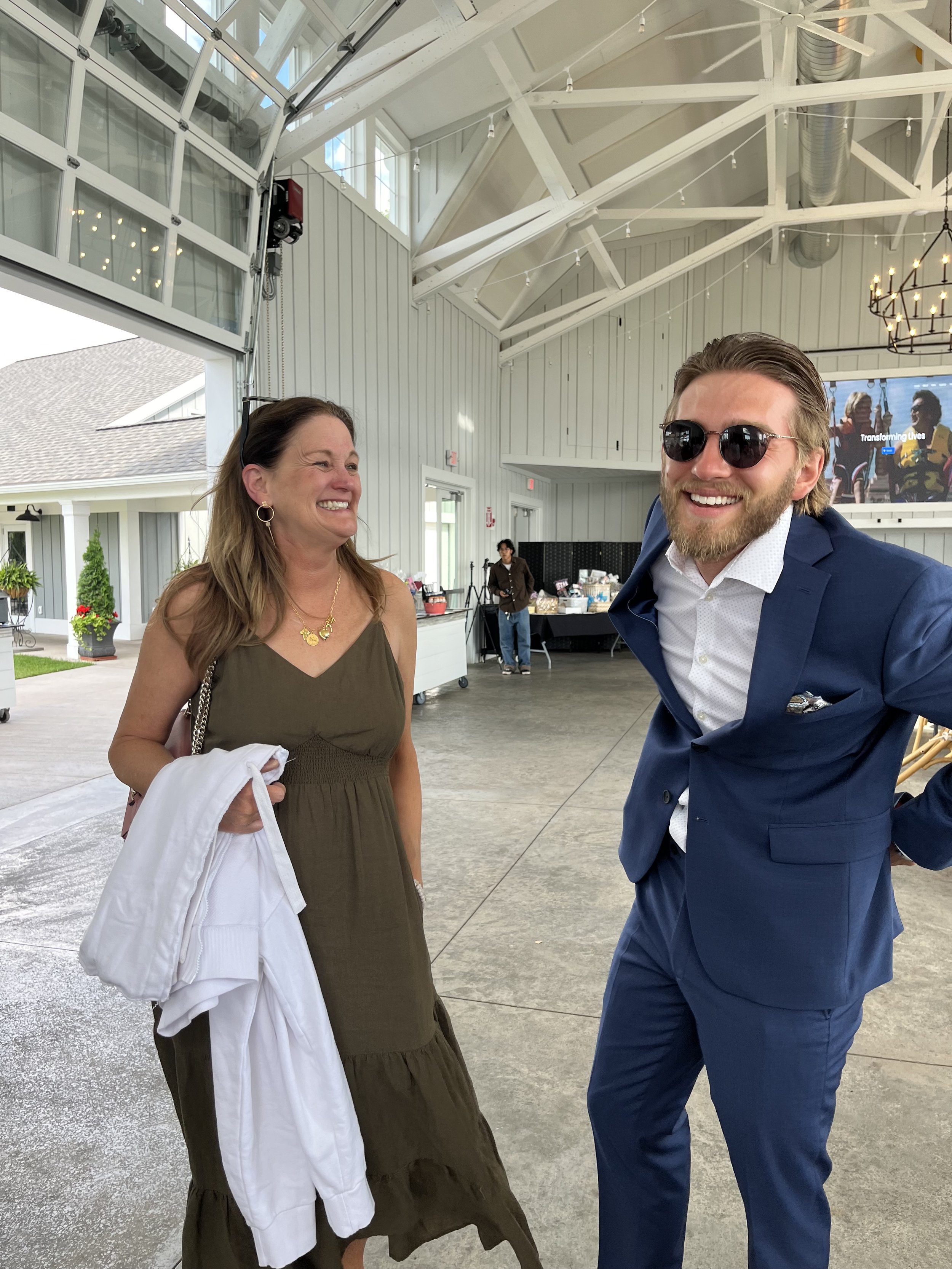 A man in a blue suit and sunglasses smiling, standing next to a woman in a brown dress holding a white jacket in a spacious indoor setting.