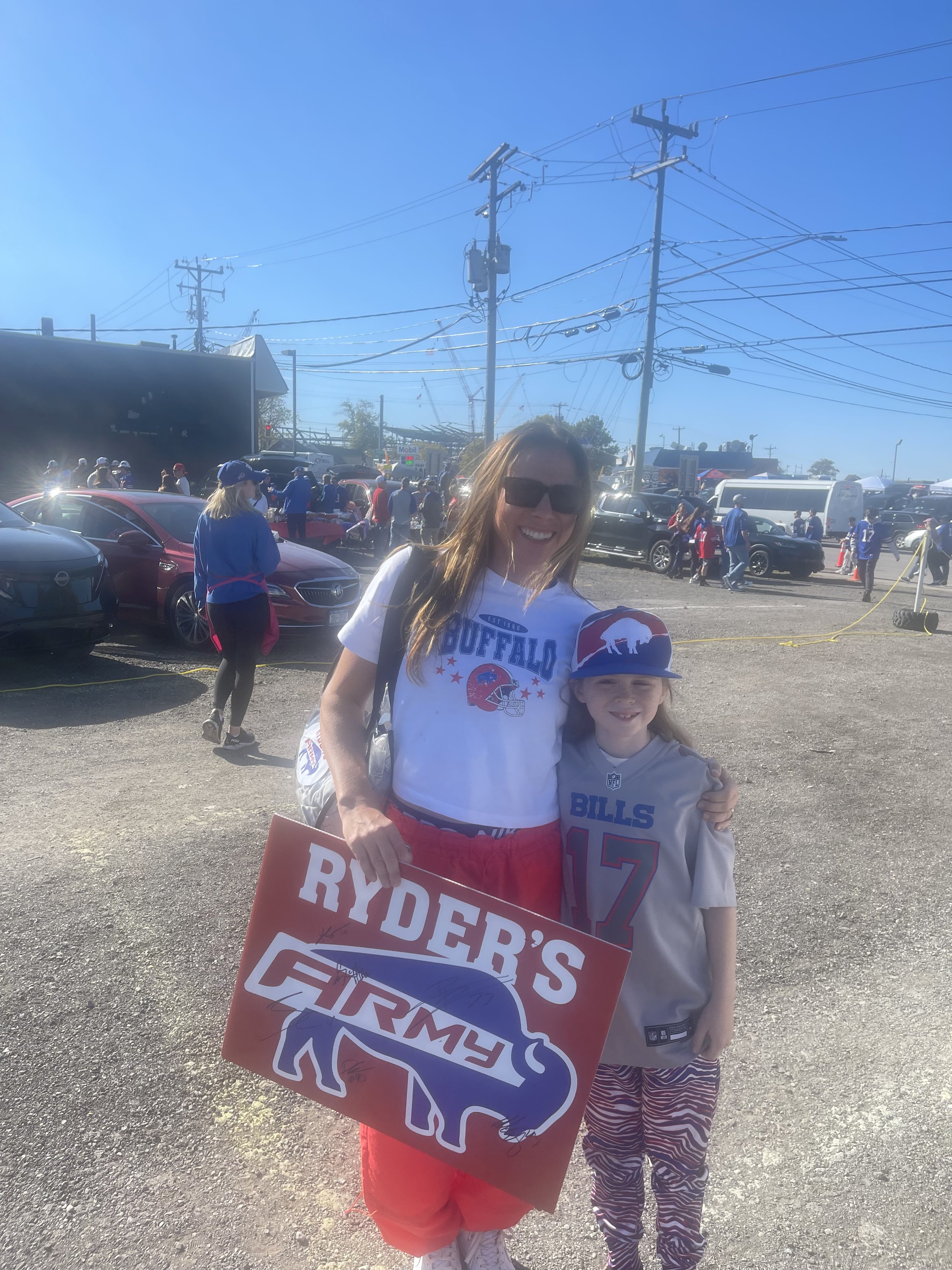 Two people wearing sports fan attire, holding a sign with text "Ryder's Firmy" and a buffalo image, standing in a parking lot with cars and other people in the background.