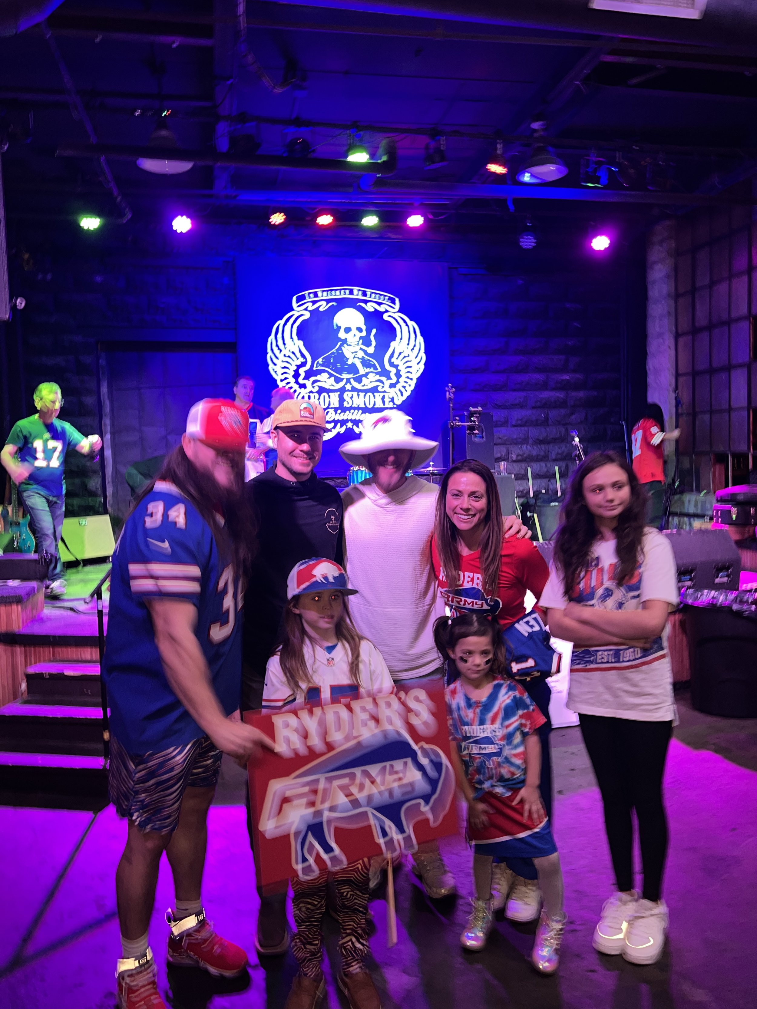 Group of people at a live music venue with Buffalo Bills merchandise and a "Ryder's Firm" sign.