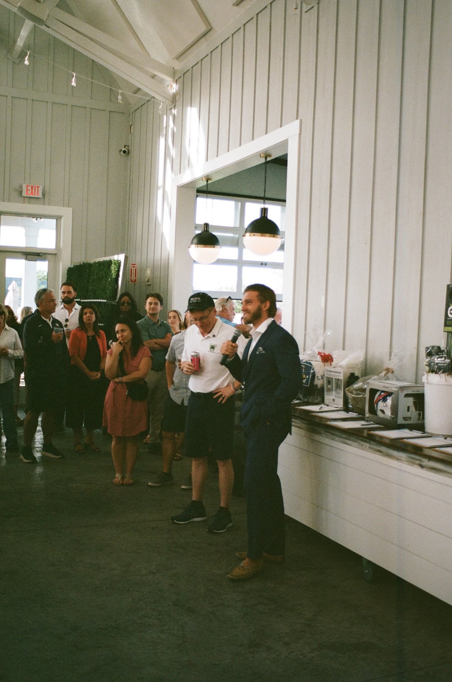 A man in a suit speaking to a group inside a spacious, well-lit room with tall ceilings. People are casually dressed and standing around listening. There's a table with raffle items, such as helmets and gift baskets, at the side.