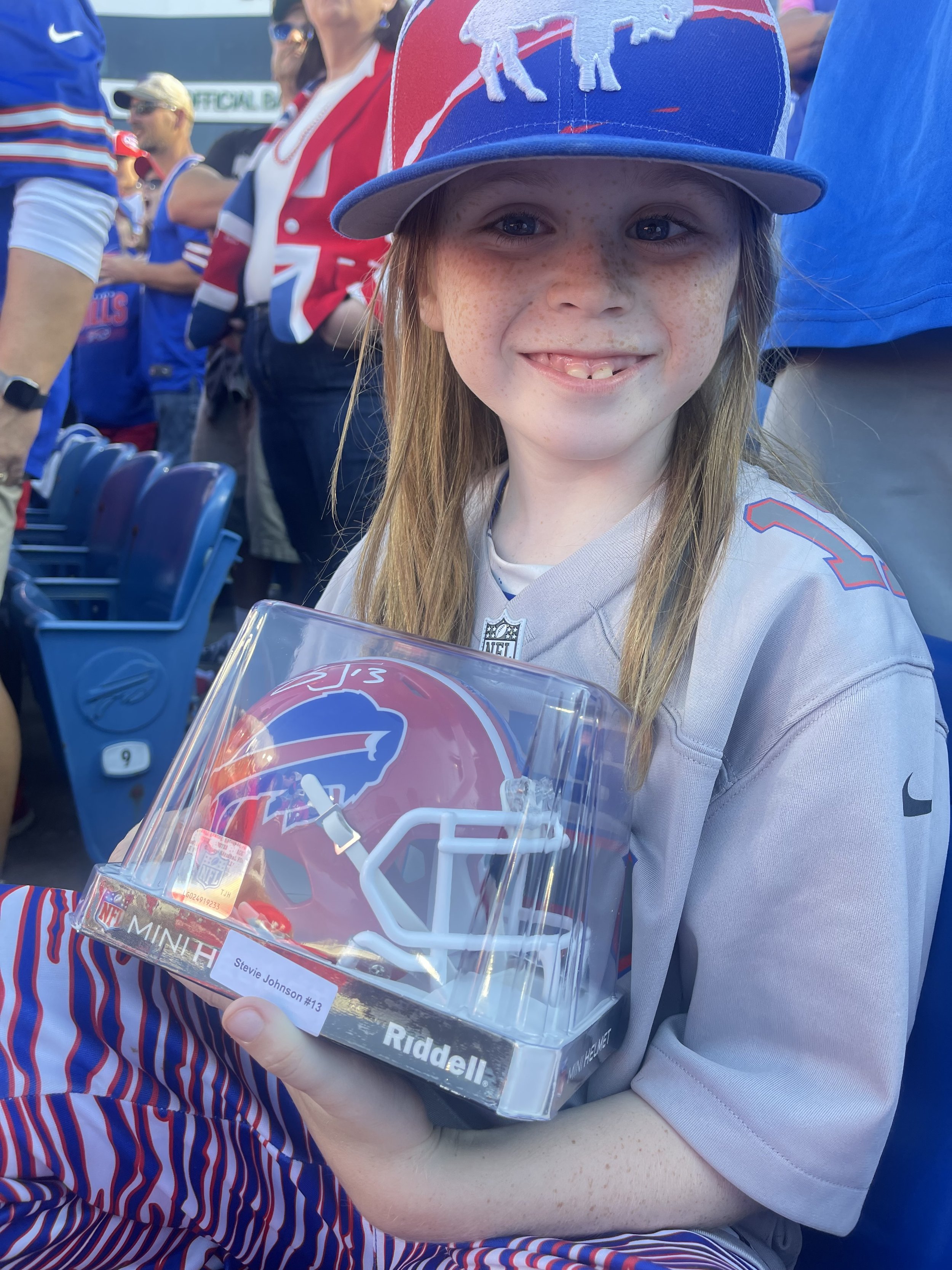 A young child wearing a Buffalo Bills cap and jersey smiles while holding a miniature Buffalo Bills helmet at a sports event. The background shows other fans in similar team apparel.