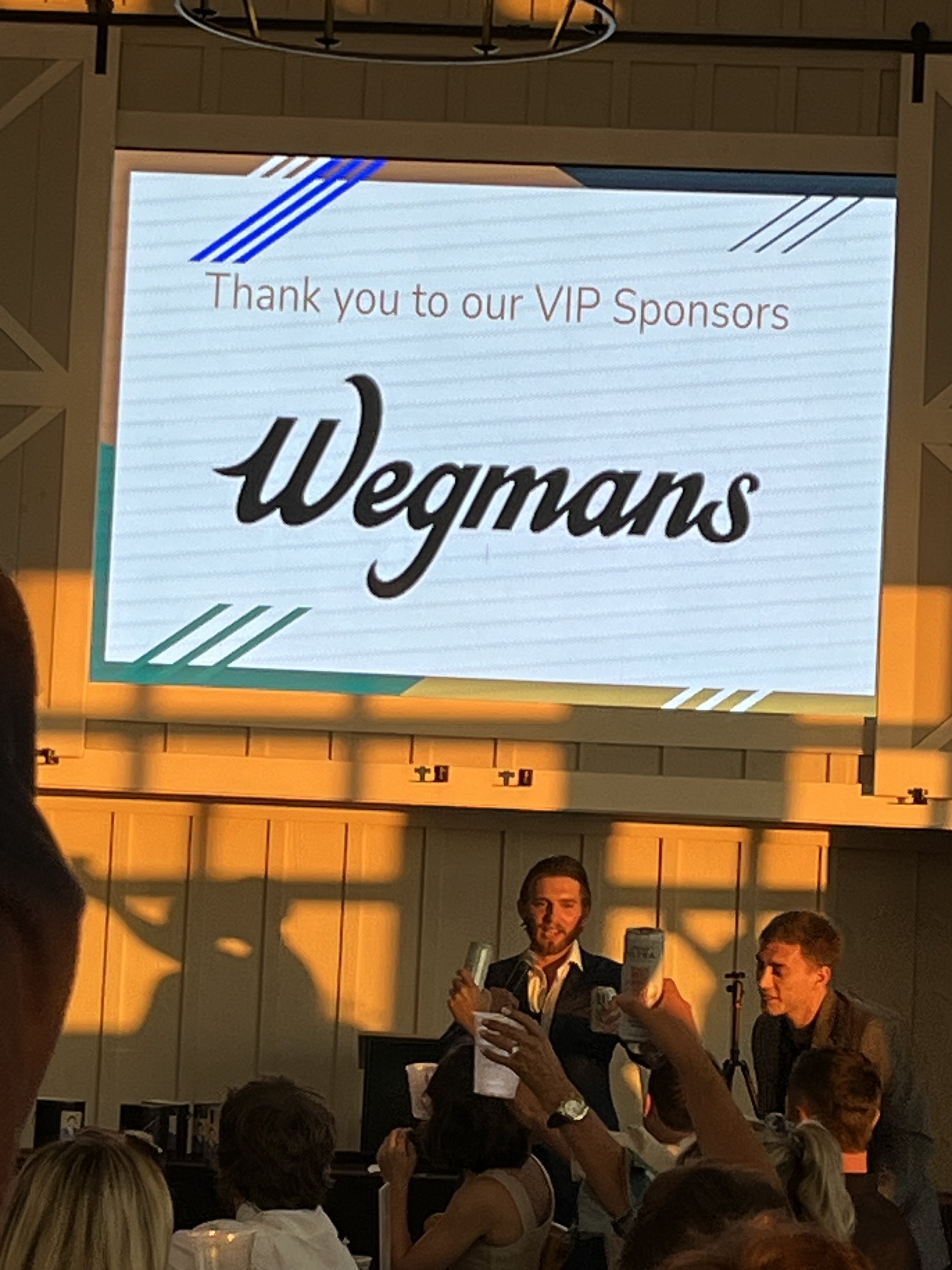 Event with people raising drinks, screen displaying 'Thank you to our VIP Sponsors Wegmans.'