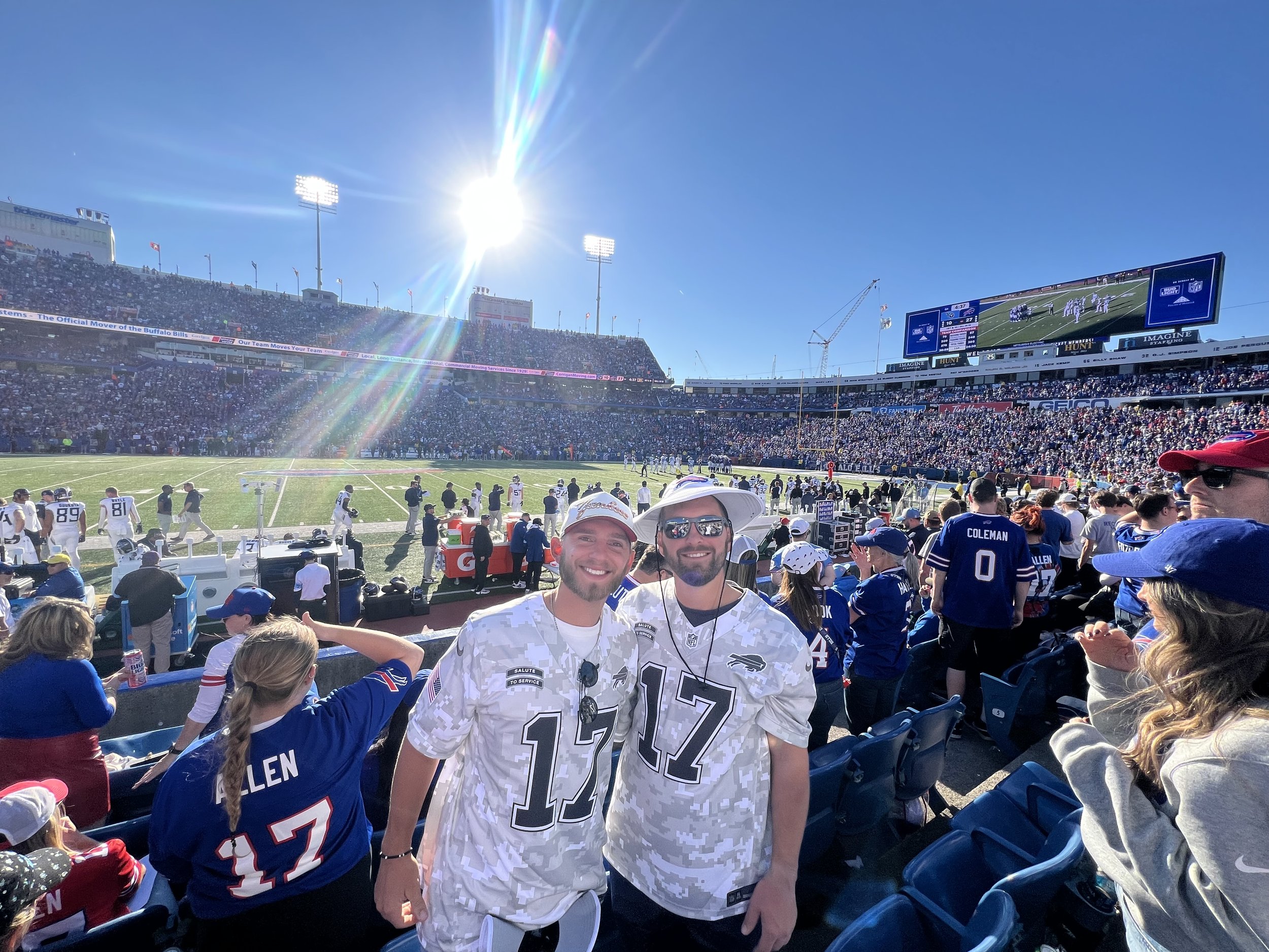 Football stadium with fans, men wearing number 17 jerseys in foreground, sunny day, large crowd in background, teams on field.