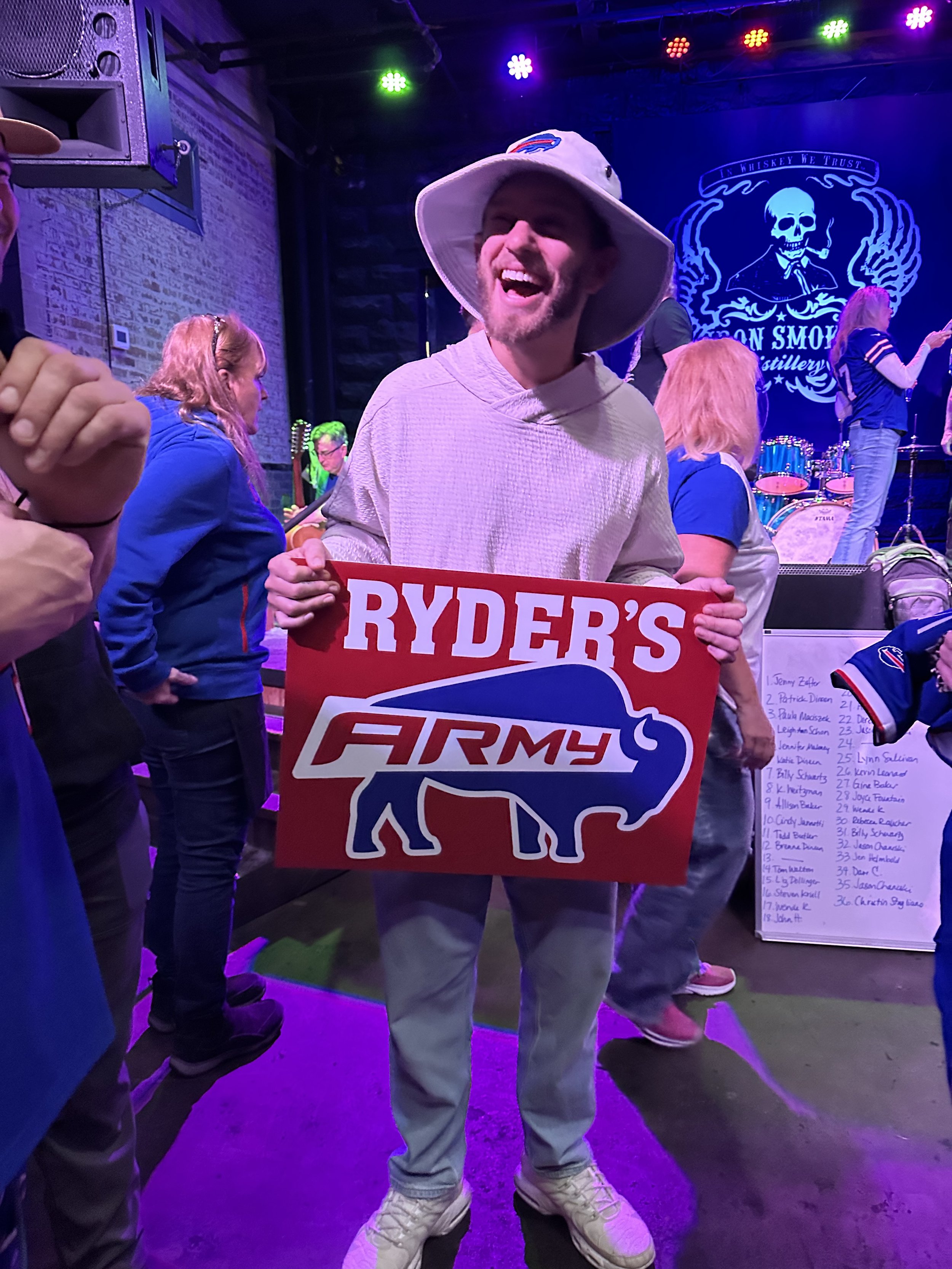 A person in a light-colored hoodie and bucket hat holds a "Ryder's Army" sign with a buffalo graphic, in a lively indoor setting with purple stage lights and people in sports jerseys.